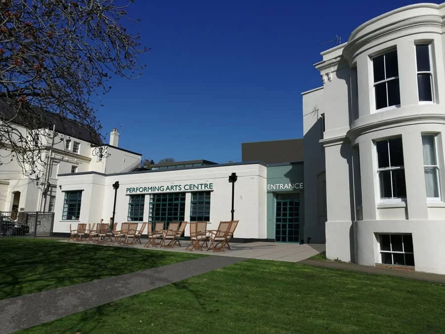 White building with “Performing Arts Centre” sign, deck chairs outside, and a green lawn under a clear blue sky.