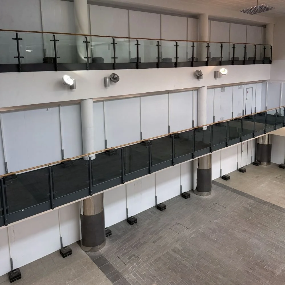 A glass-fenced walkway leads to blocked doors on the upper floor of an atrium with white walls and gray tile flooring.