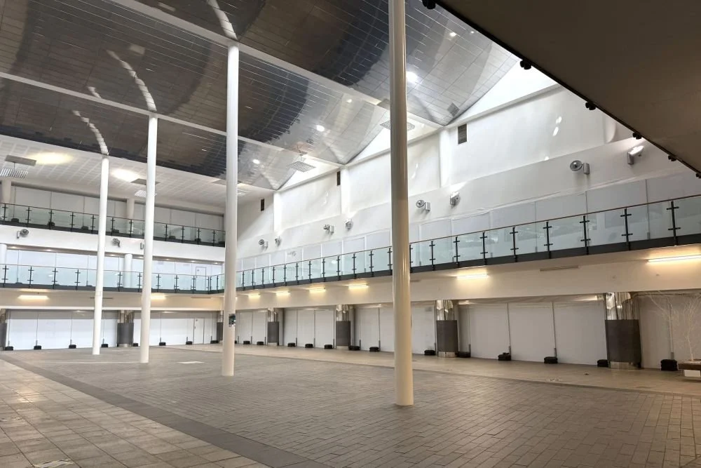 Spacious, empty indoor hall with high ceilings, skylights, and a second-floor glass railing balcony surrounding the open area.