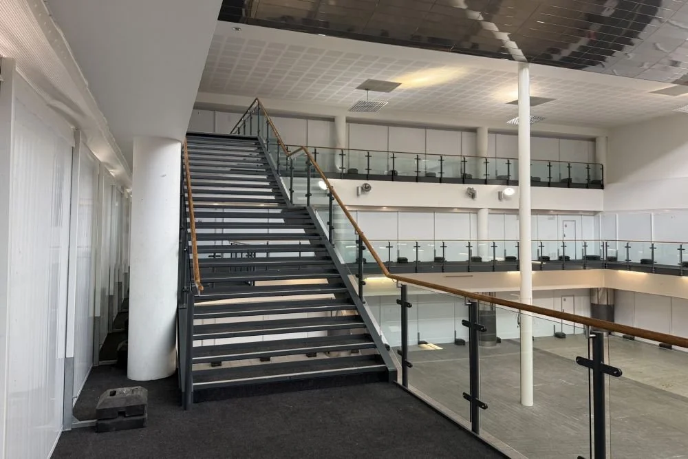 A modern, empty office interior with a wide staircase and glass railings leading to an upper mezzanine level.