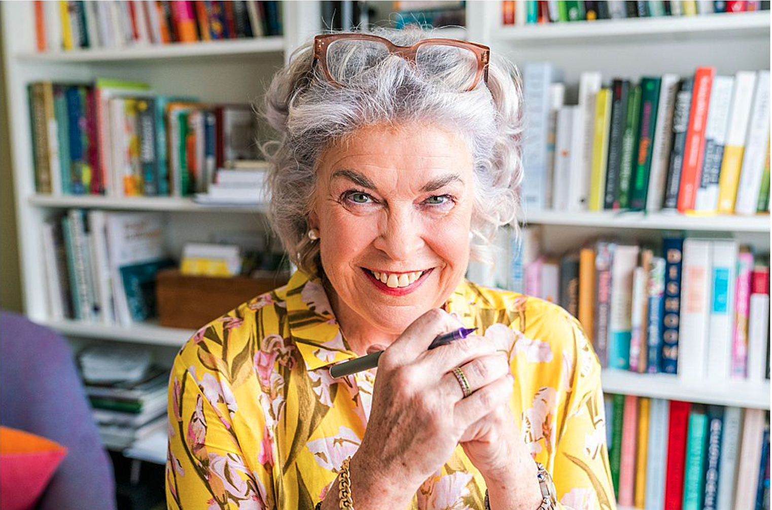An elderly woman with gray curly hair and glasses on her head, smiling while holding a pen, sitting in front of a bookshelf filled with colorful books.