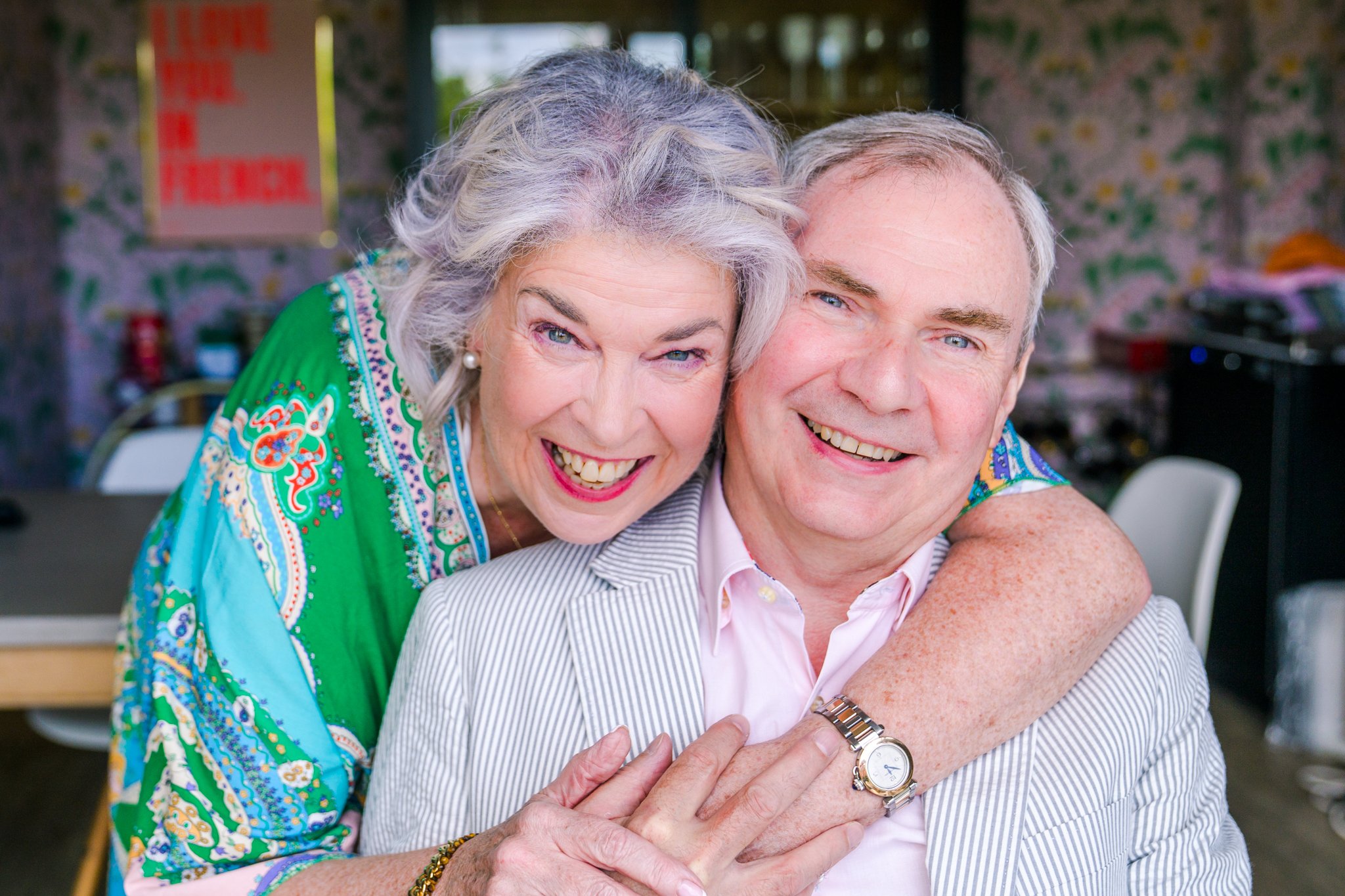 A smiling elderly woman with gray hair hugging a smiling elderly man with gray hair. The woman is wearing a colorful, patterned shirt and pearl earrings, and the man is wearing a striped blazer and a watch. They are in an indoor setting with a floral-patterned backdrop.