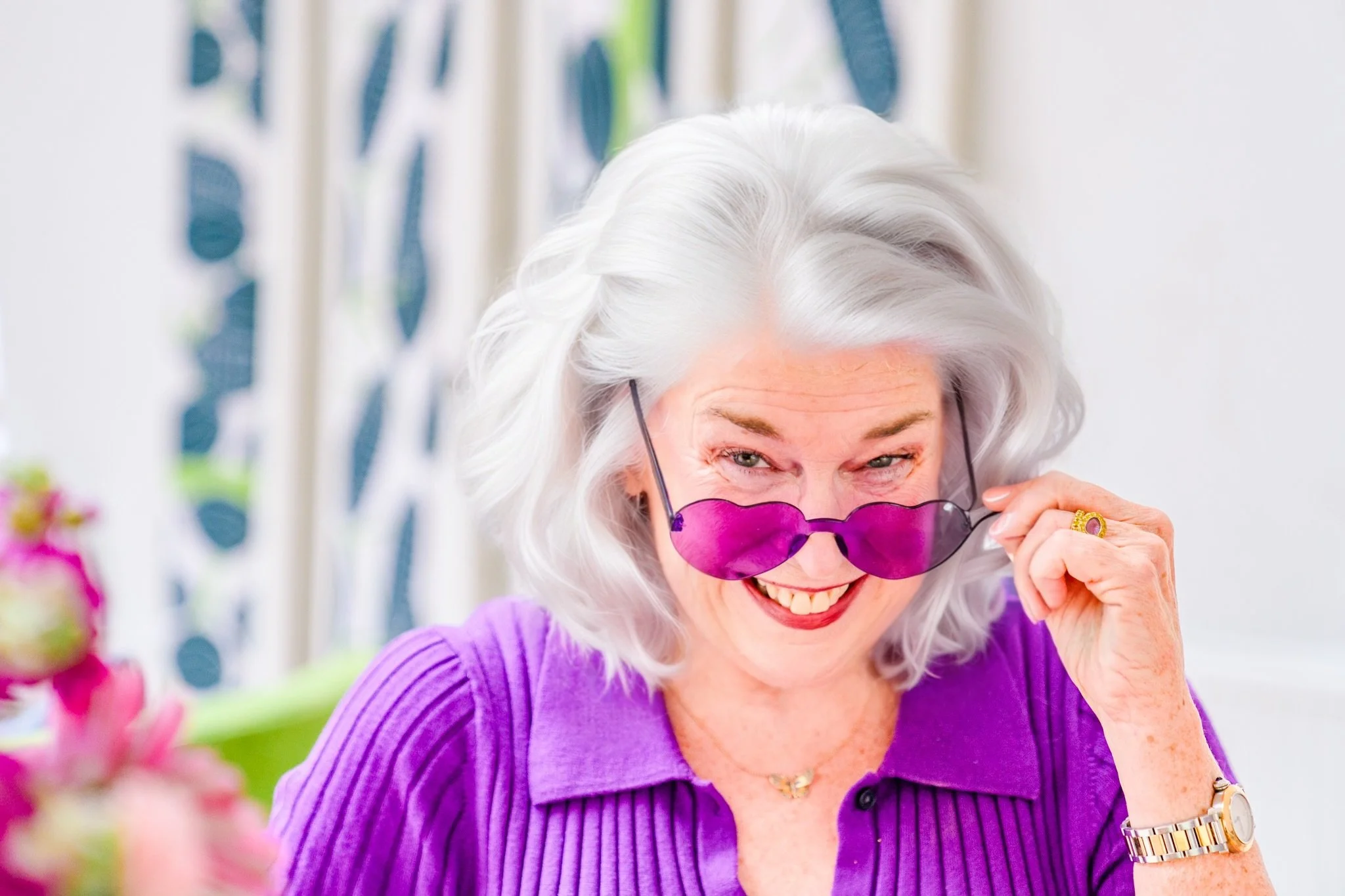A smiling senior woman with white hair, wearing purple sunglasses and a purple top, holding the sunglasses with one hand, in a brightly lit indoor space with blurred background.