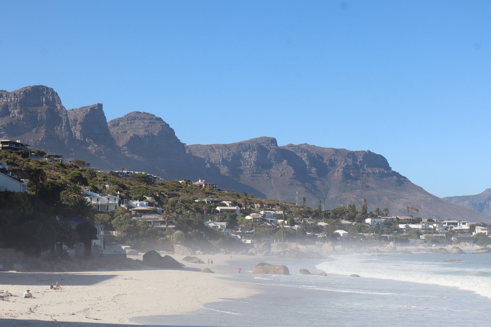 Sunny beach with white sand and large rocks, houses on a hillside, and a mountain range in the background