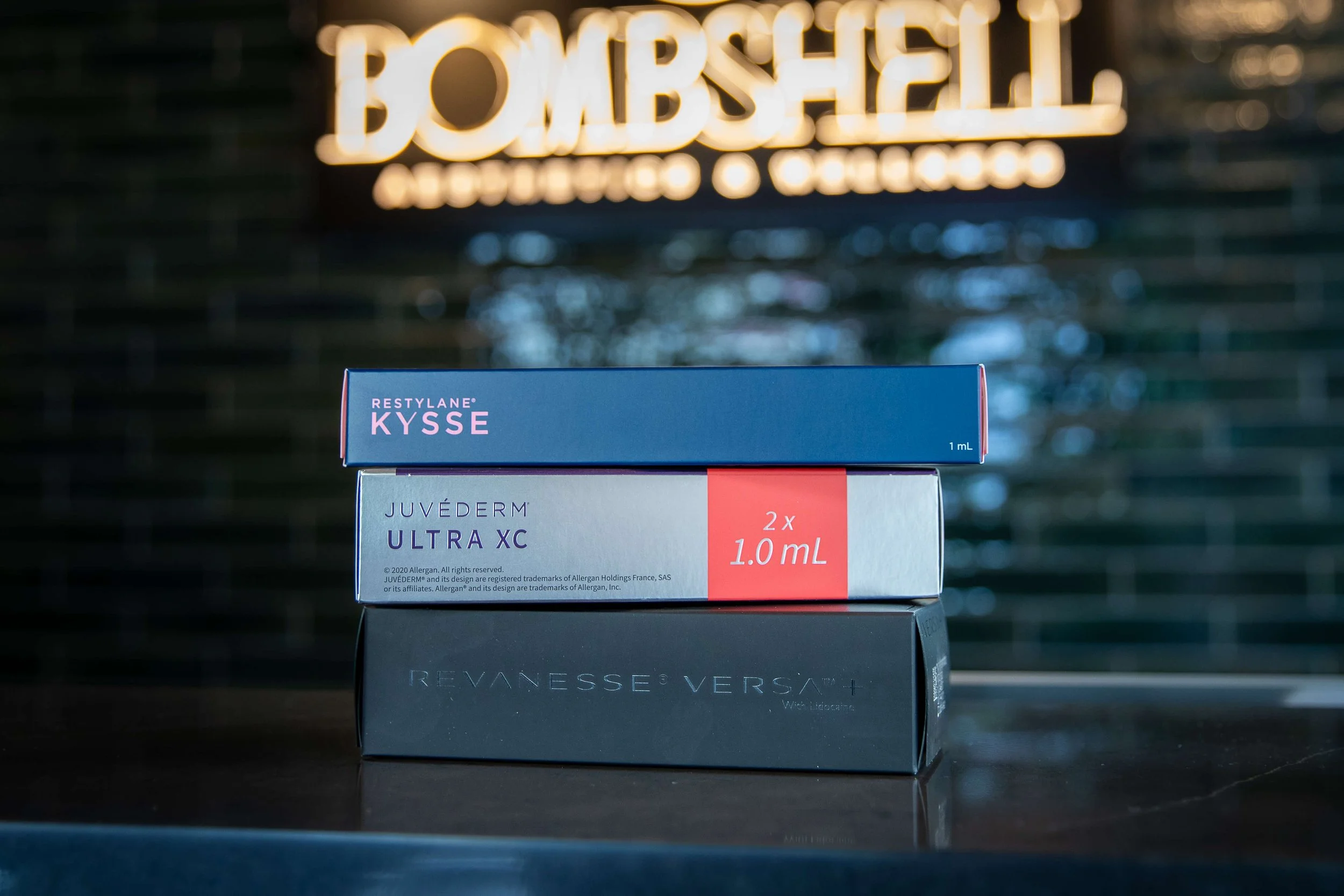 Stacked dermal filler product boxes displayed on a counter in a medical aesthetics setting, with Bombshell Aesthetics signage softly blurred in the background.