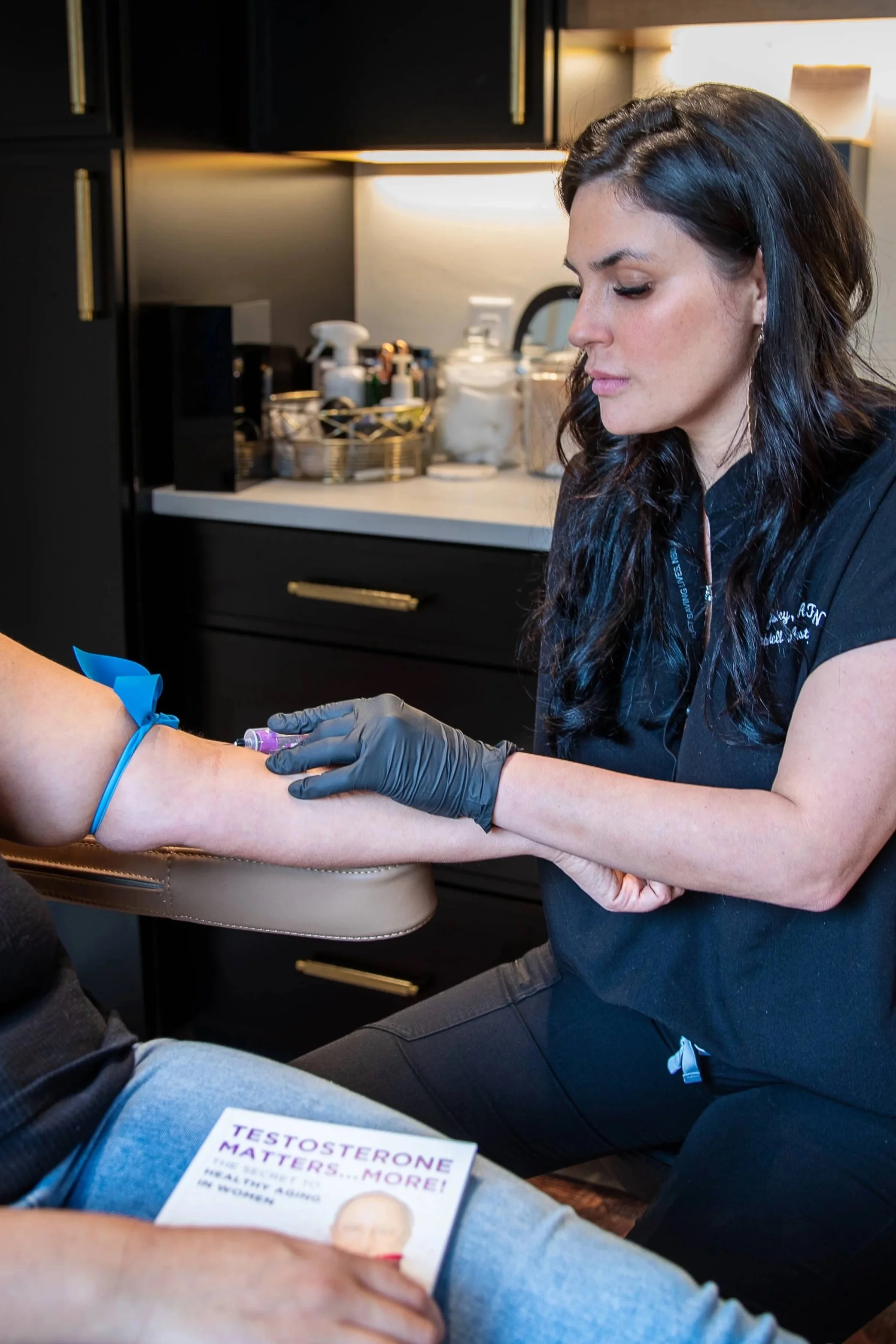 Provider at Bombshell Aesthetics drawing blood for hormone replacement therapy (HRT) in a modern treatment room, while the seated patient holds a copy of the book “Testosterone Matters… More!” on their lap.