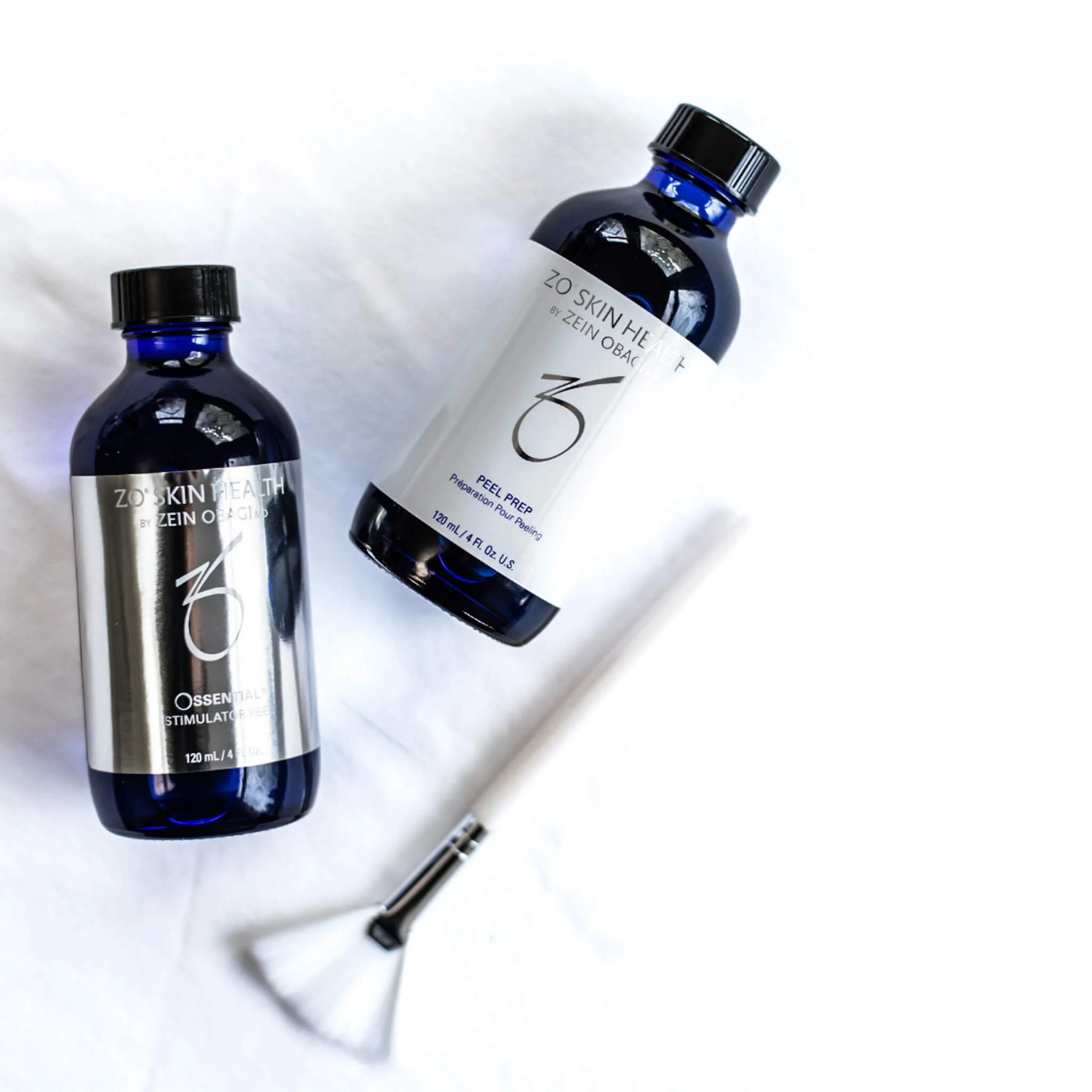 Two ZO Skin Health bottles—Ossential Stimulator Peel and Peel Prep—placed on a light marble surface with a small fan brush nearby, styled in a clean, professional skincare setting.