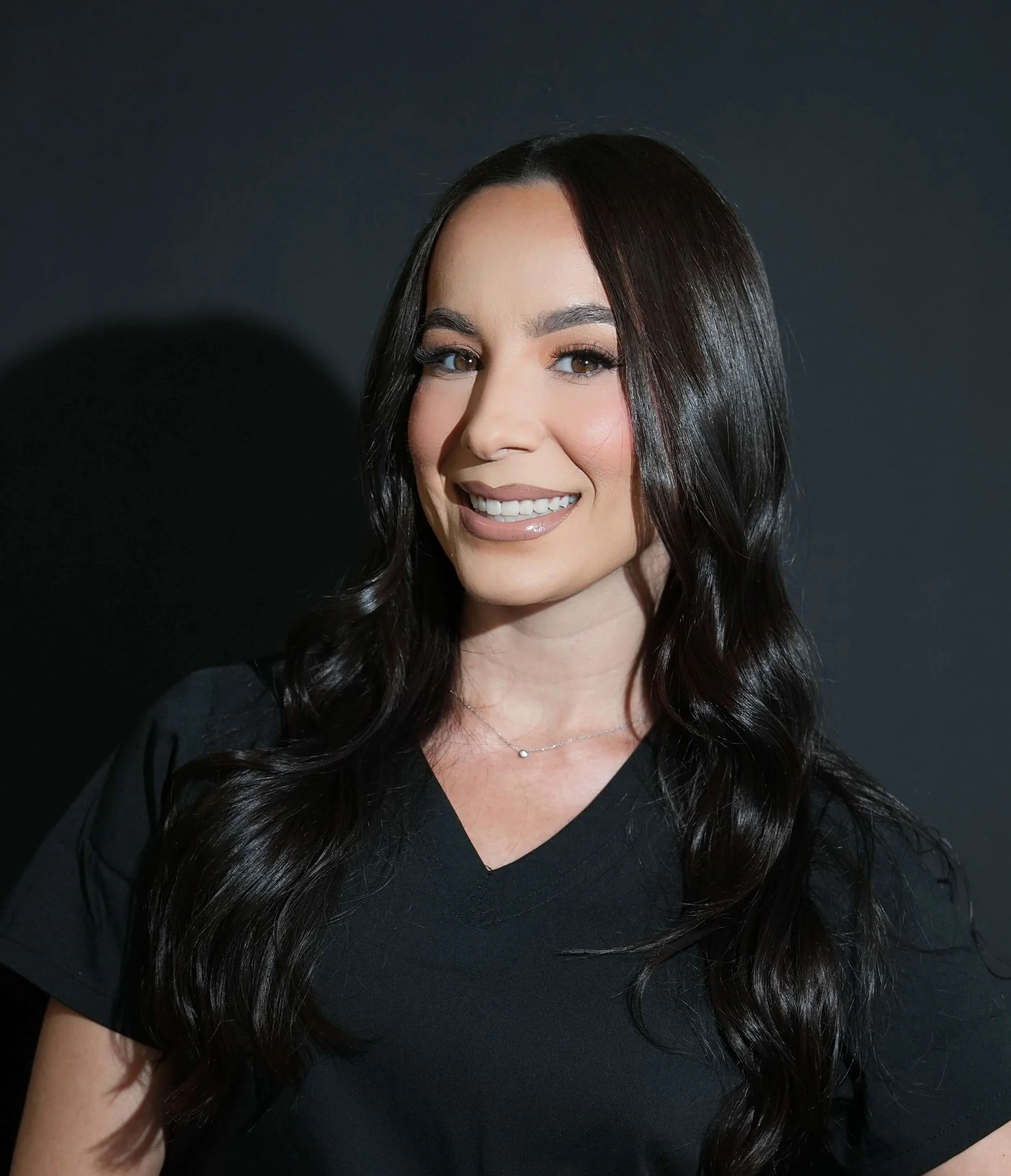 Close-up portrait of a smiling woman with long, dark, wavy hair against a dark background at Bombshell Aesthetics in Bedford, NH