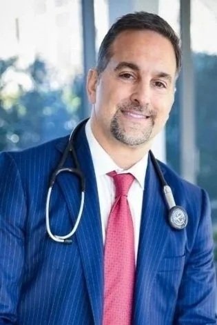 A male doctor wearing a blue suit and pink tie, with a stethoscope around his neck, smiling in front of a window.