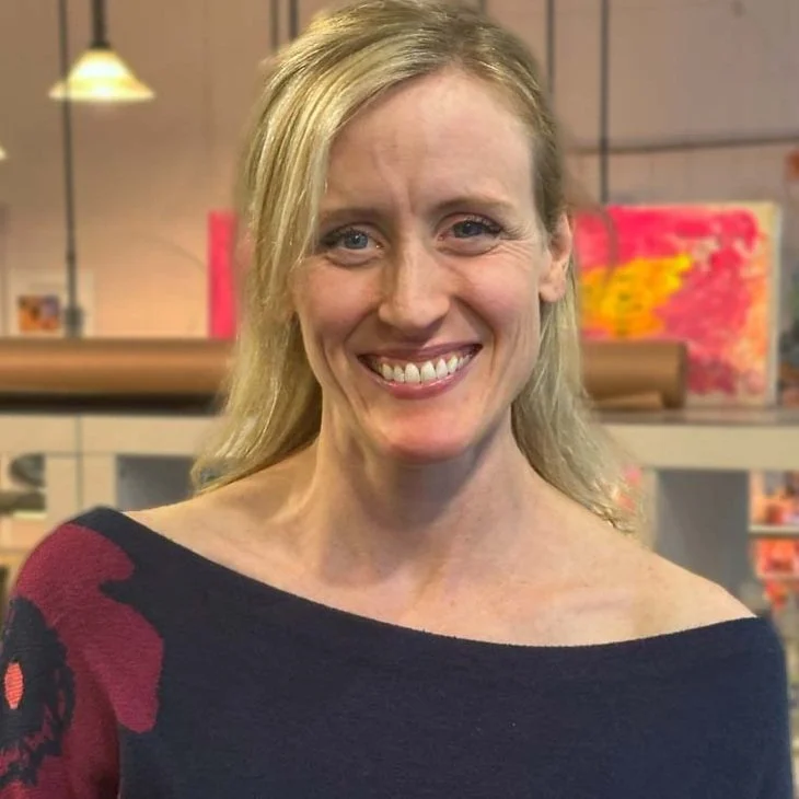 A woman with blonde hair, smiling, wearing a dark top with red and black patterns, standing in a cozy indoor setting with warm lighting and colorful wall decorations.