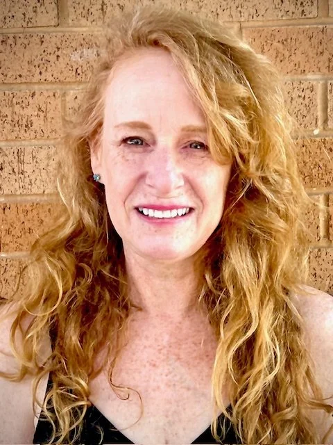 A woman with curly red hair smiling in front of a brick wall.