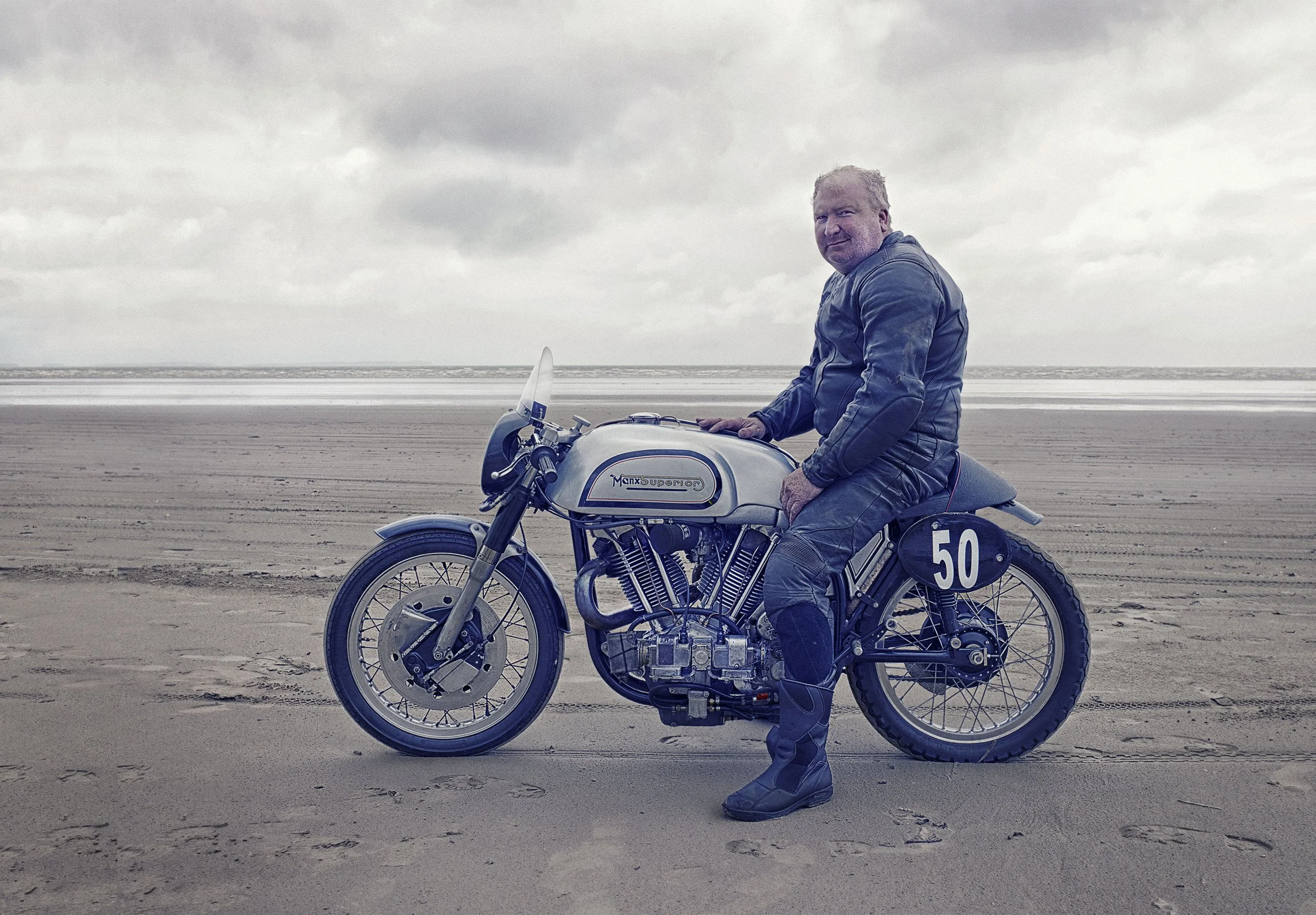 A man in motorcycle gear sitting on a vintage motorcycle on a beach with cloudy sky and ocean in the background,  Pendine Sands Land speed, Advertising Photographer, Lifestyle.  