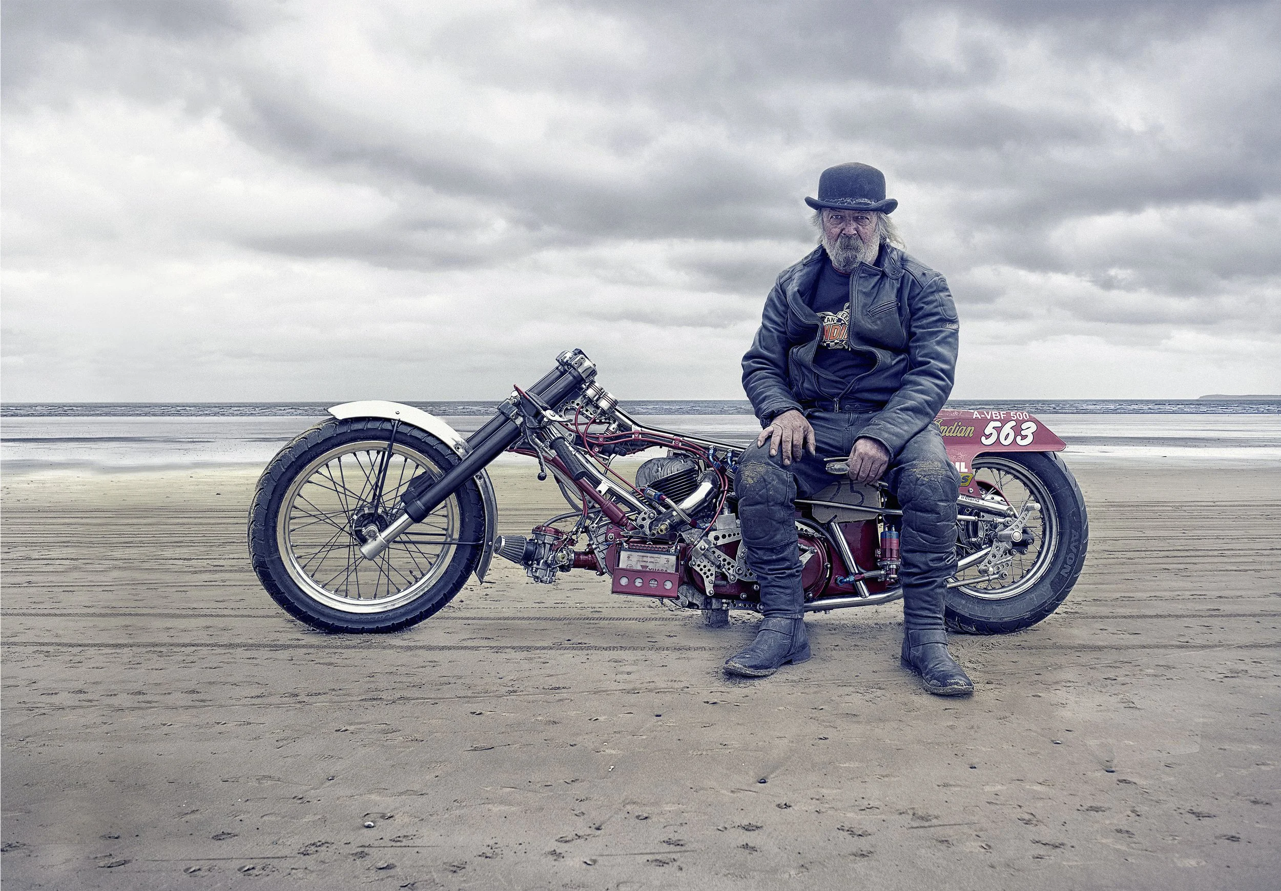 A man with gray hair and beard wearing a black hat, black jacket, and black boots sits on a vintage motorcycle on a sandy beach under cloudy sky.