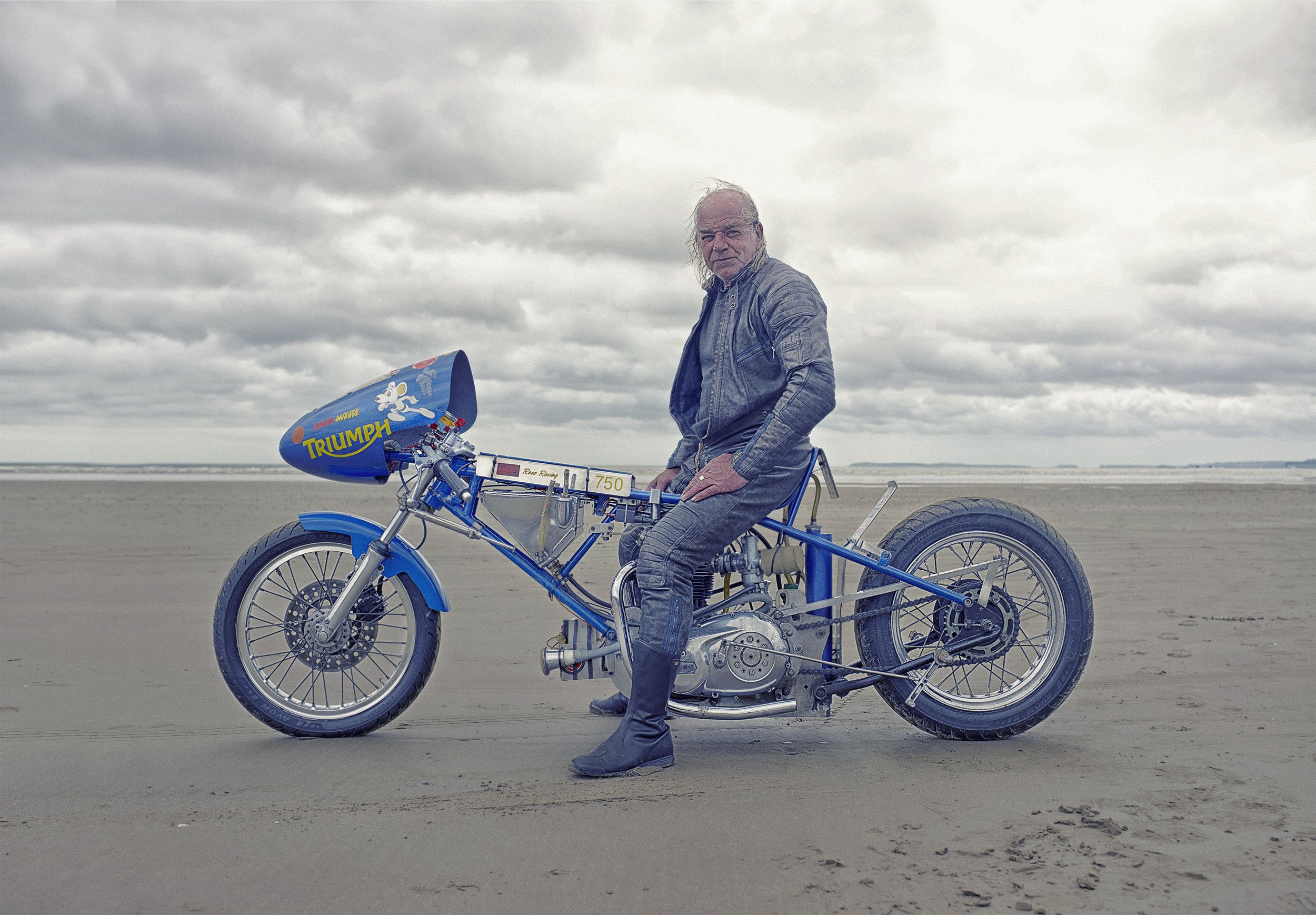 A man sitting on a vintage motorcycle with a Triumph logo, on a sandy beach with overcast sky in the background.