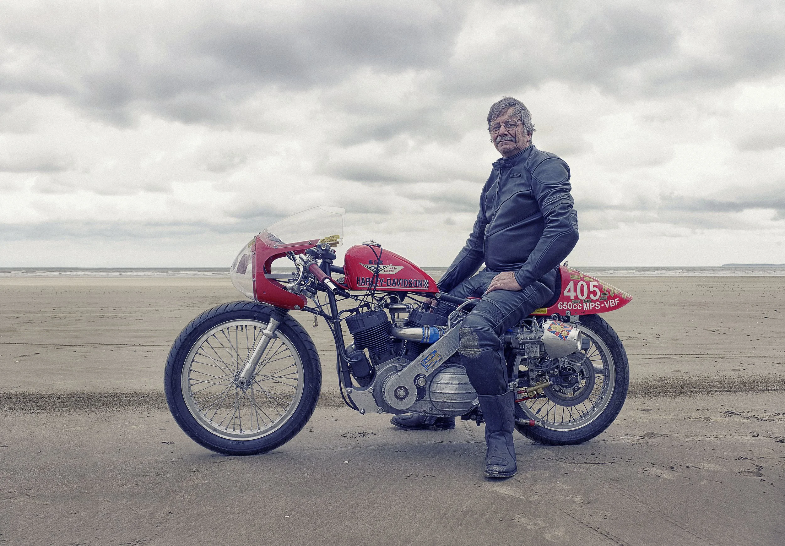 Man sitting on a vintage Harley-Davidson racing motorcycle on a sandy beach with cloudy sky in the background Pendine Sands Land speed,Pendine Sands Land speed, Advertising Photographer, Lifestyle.   