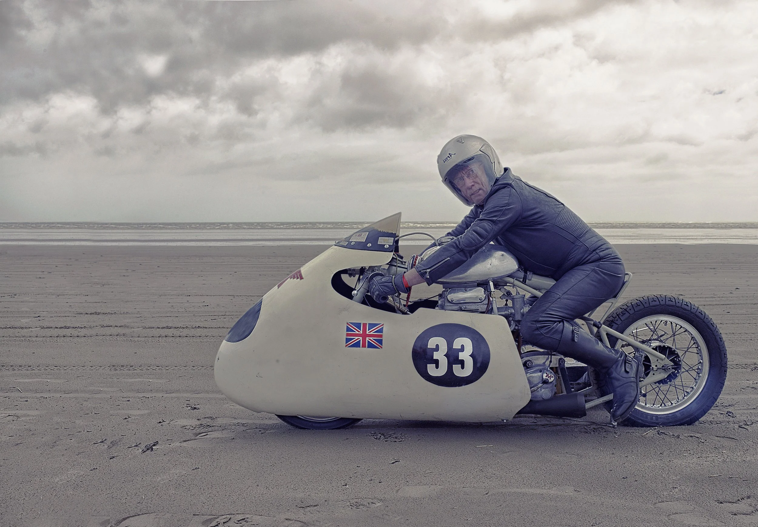 A man dressed in leather riding gear on a vintage motorcycle with a sidecar on a sandy beach under cloudy skies.