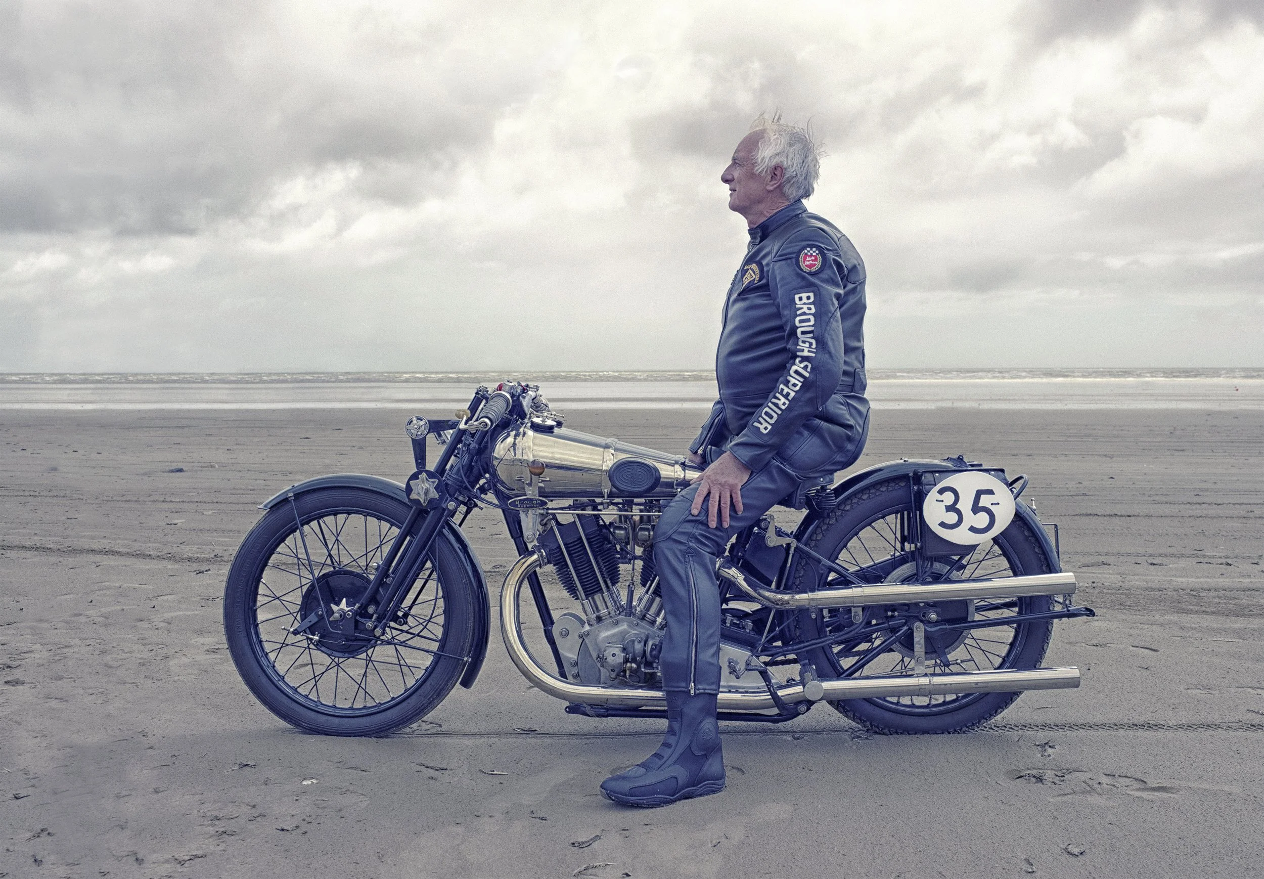 An elderly man in a vintage motorcycle racing suit sits on a classic motorcycle on a sandy beach under a cloudy sky.