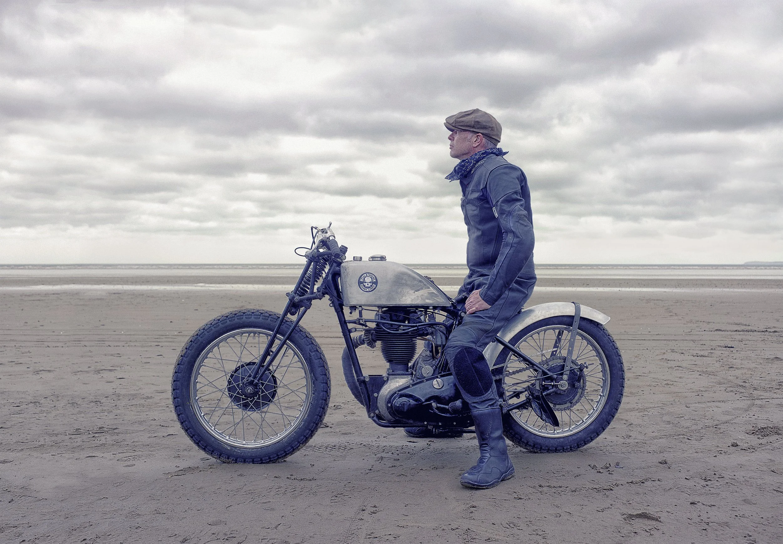A man dressed in vintage motorcycle gear sitting on a retro motorcycle on a sandy beach with overcast sky, Pendine Sands Land speed, Advertising Photographer, Lifestyle.  