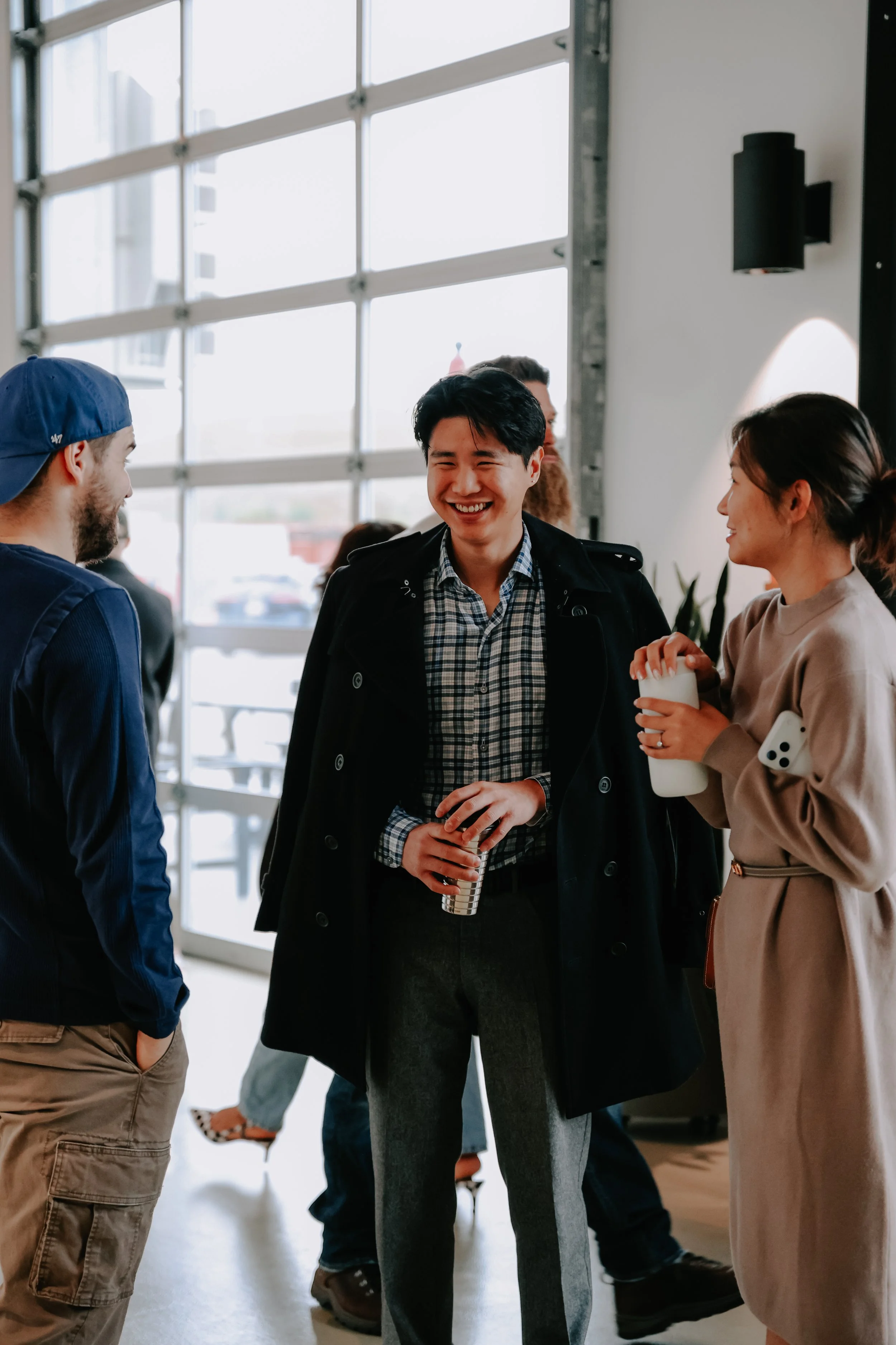 People socializing indoors near large windows, smiling and engaging in conversation.