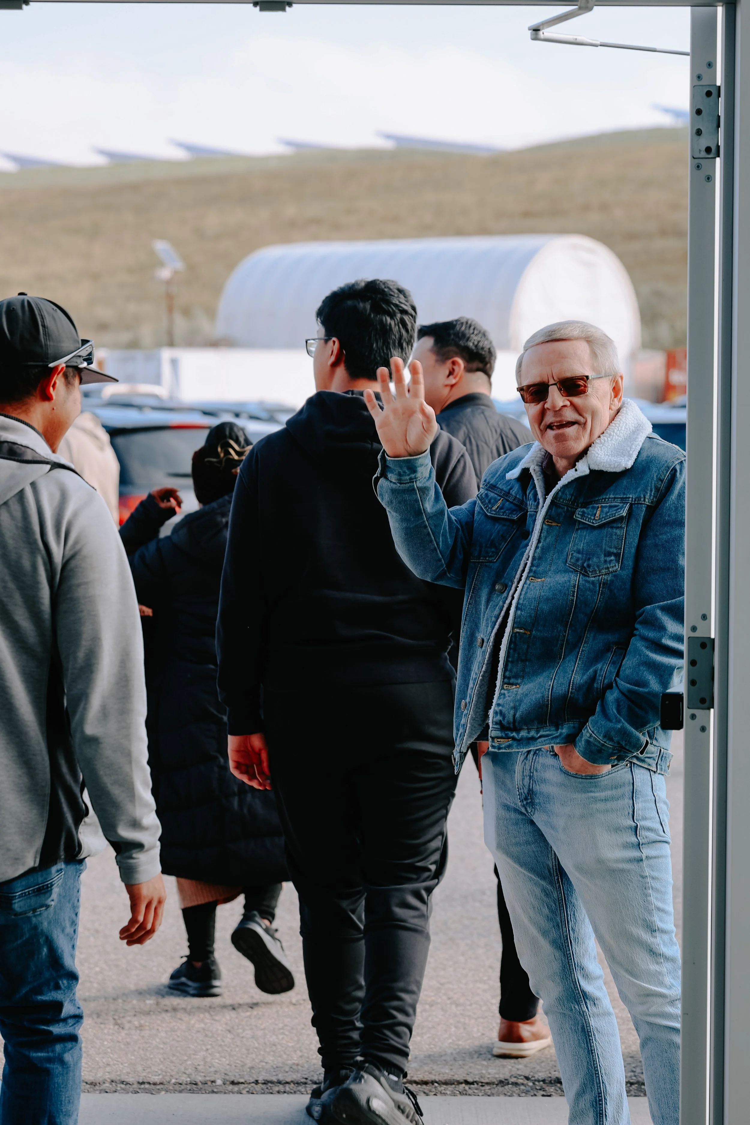 People waiting in line outdoors at an airport, with a man waving and smiling at the camera.