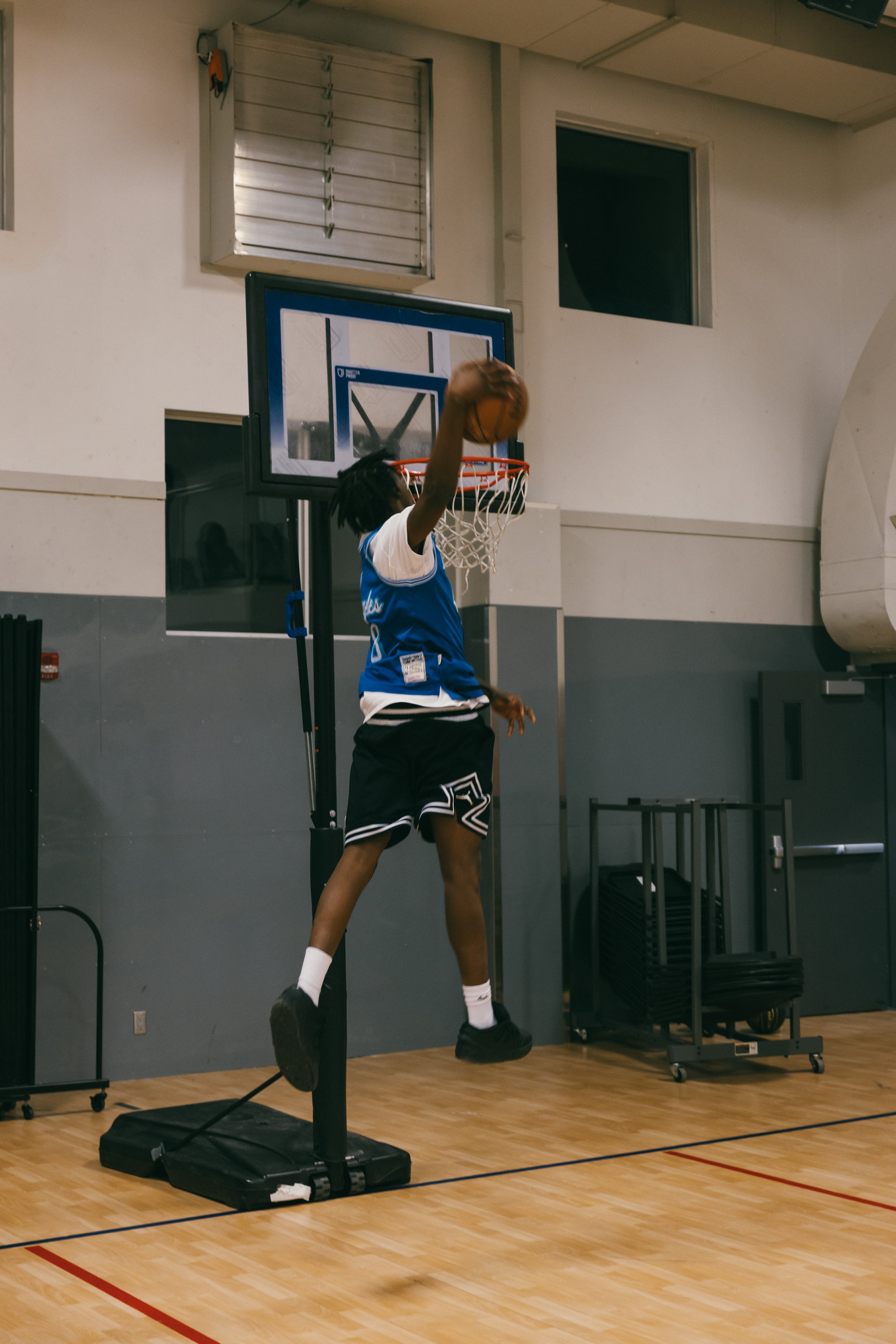 A person jumping high to make a basketball shot in an indoor gym, with basketball hoop visible behind them. The person is wearing a blue jersey, black shorts, white socks, and black shoes.