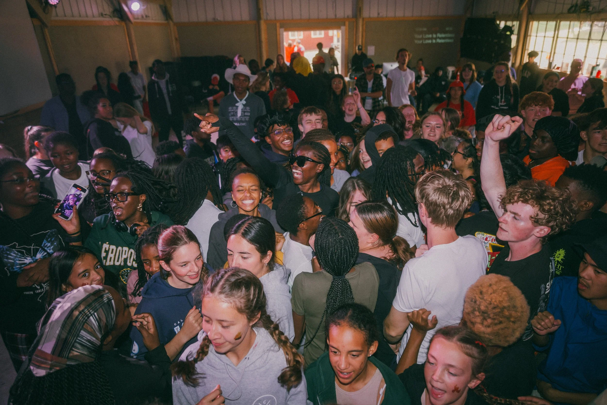 A large group of young people dancing and celebrating indoors. Some are smiling, raising fists, and taking photos.