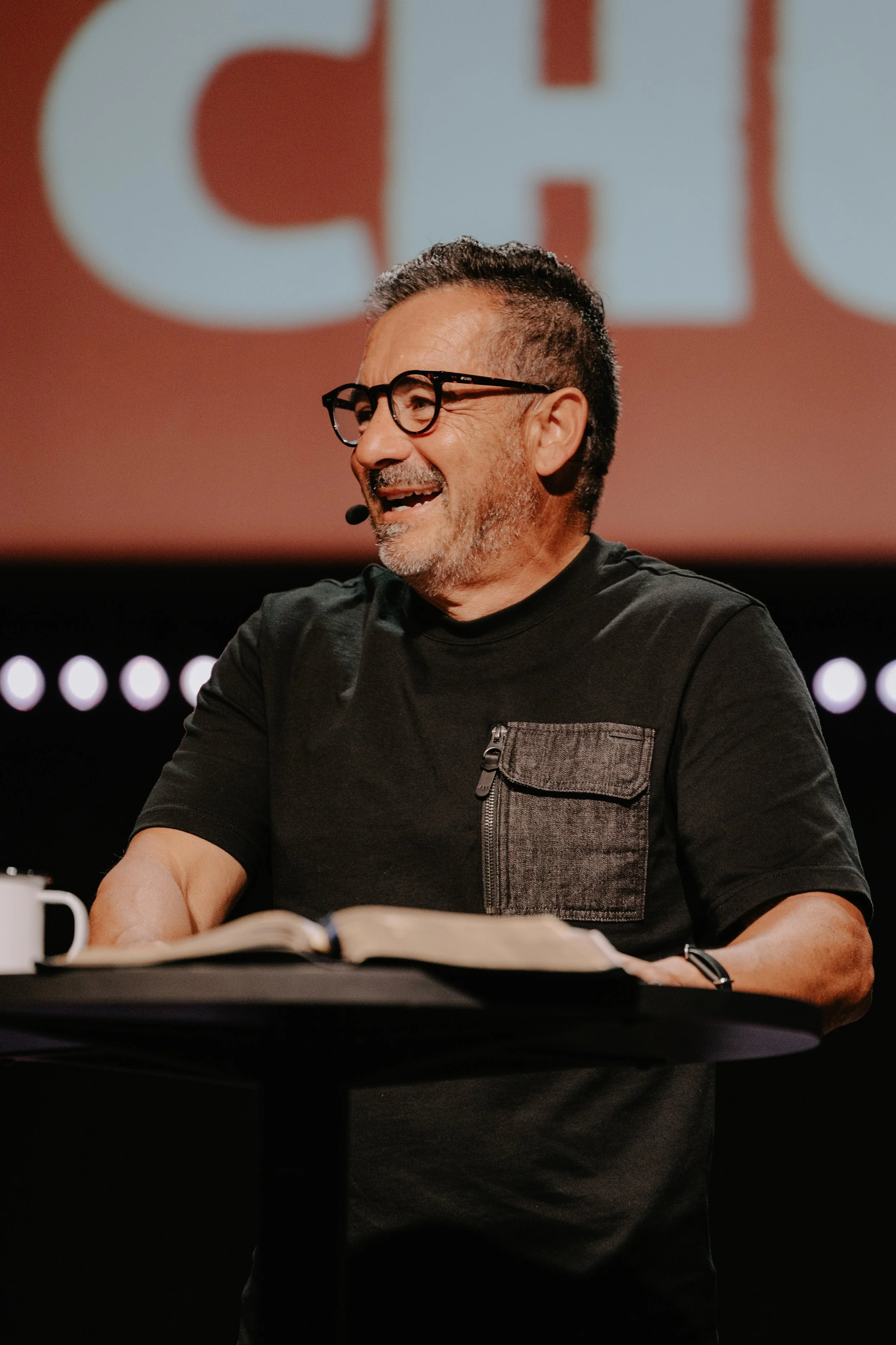 A man with glasses, a beard, and gray hair, wearing a black T-shirt with a pocket on the chest, is smiling and sitting at a table with an open book in front of him. He appears to be speaking or engaged in a conversation, with a microphone near his ch