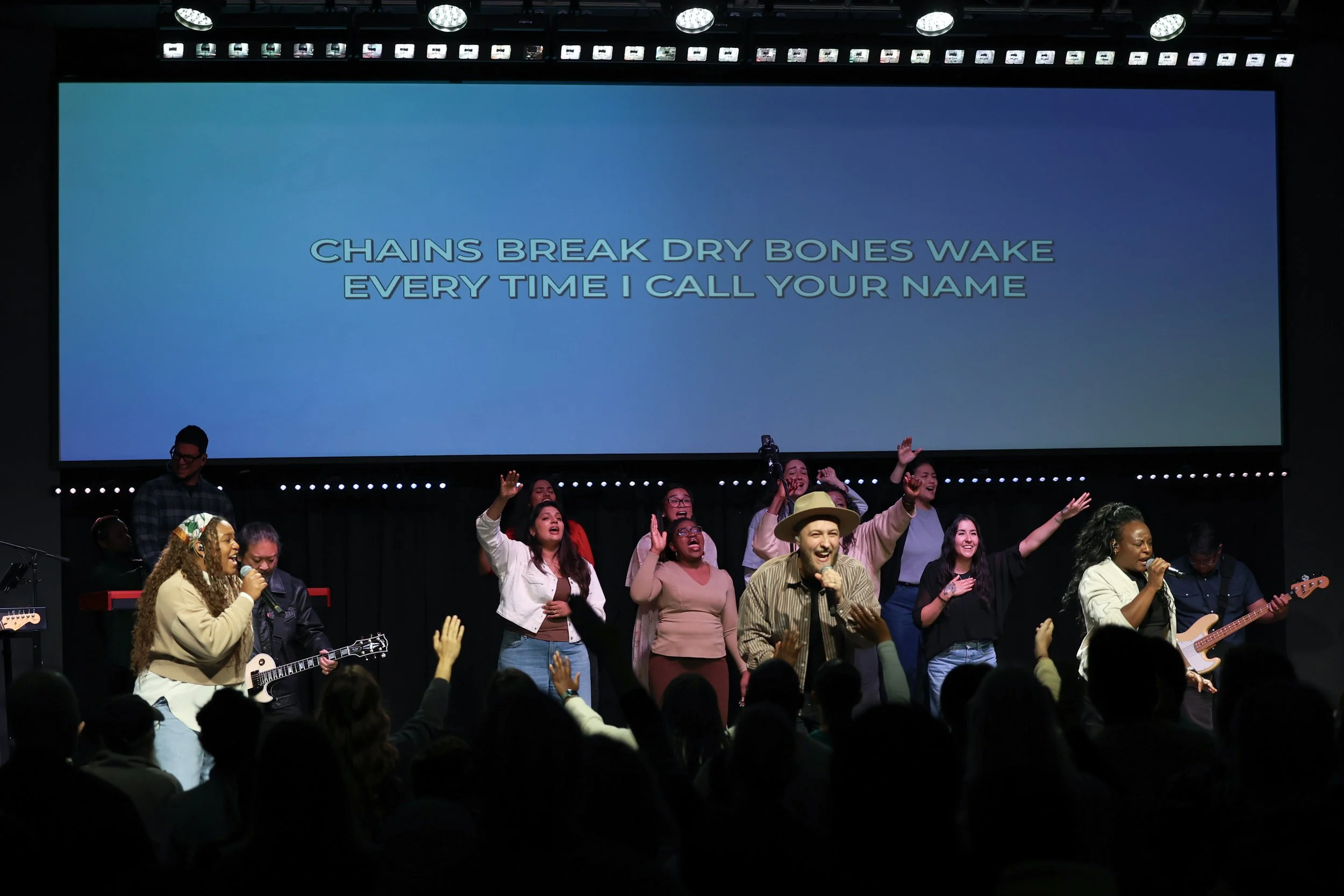 A group of people on stage performing music with a large screen behind them displaying lyrics that read, "Chains break dry bones wake every time I call your name." The audience is visible in the foreground with hands raised.