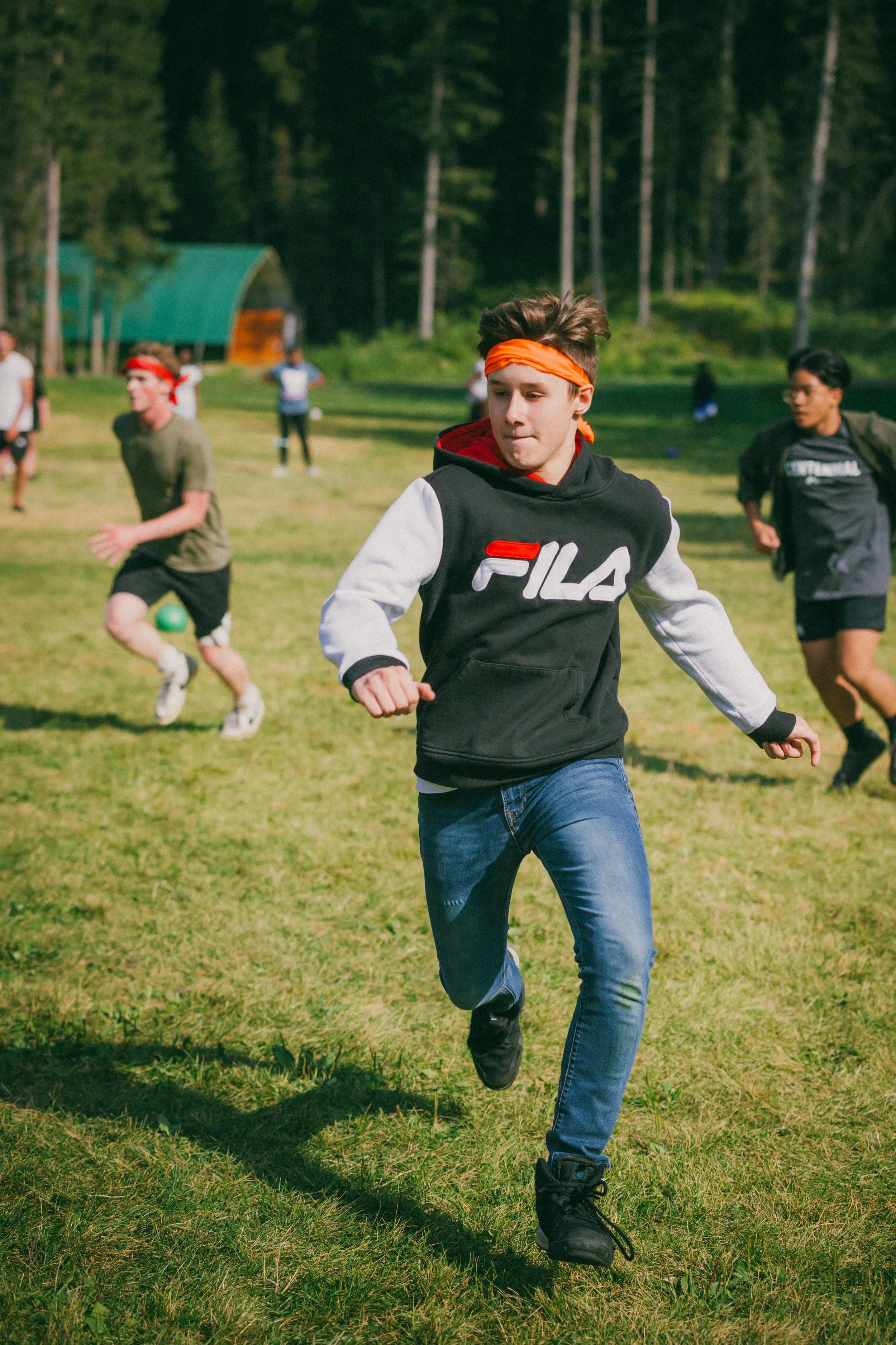 Young boy with brown hair wearing an orange headband, a black and white FILA hoodie, blue jeans, and black shoes running outdoors on a grassy field. In the background, other kids are playing, with trees and a small building visible.