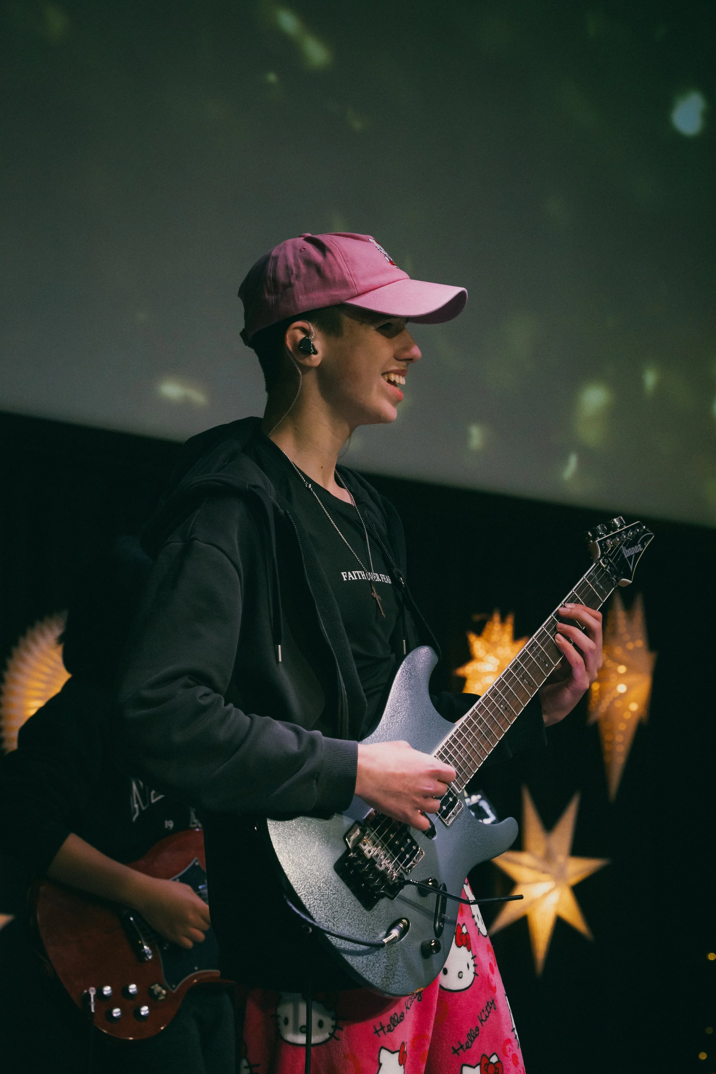A young person smiling and playing an electric guitar, wearing a pink cap, black hoodie, and Hello Kitty themed pink sweats, in a dimly lit setting with star-shaped lights in the background.