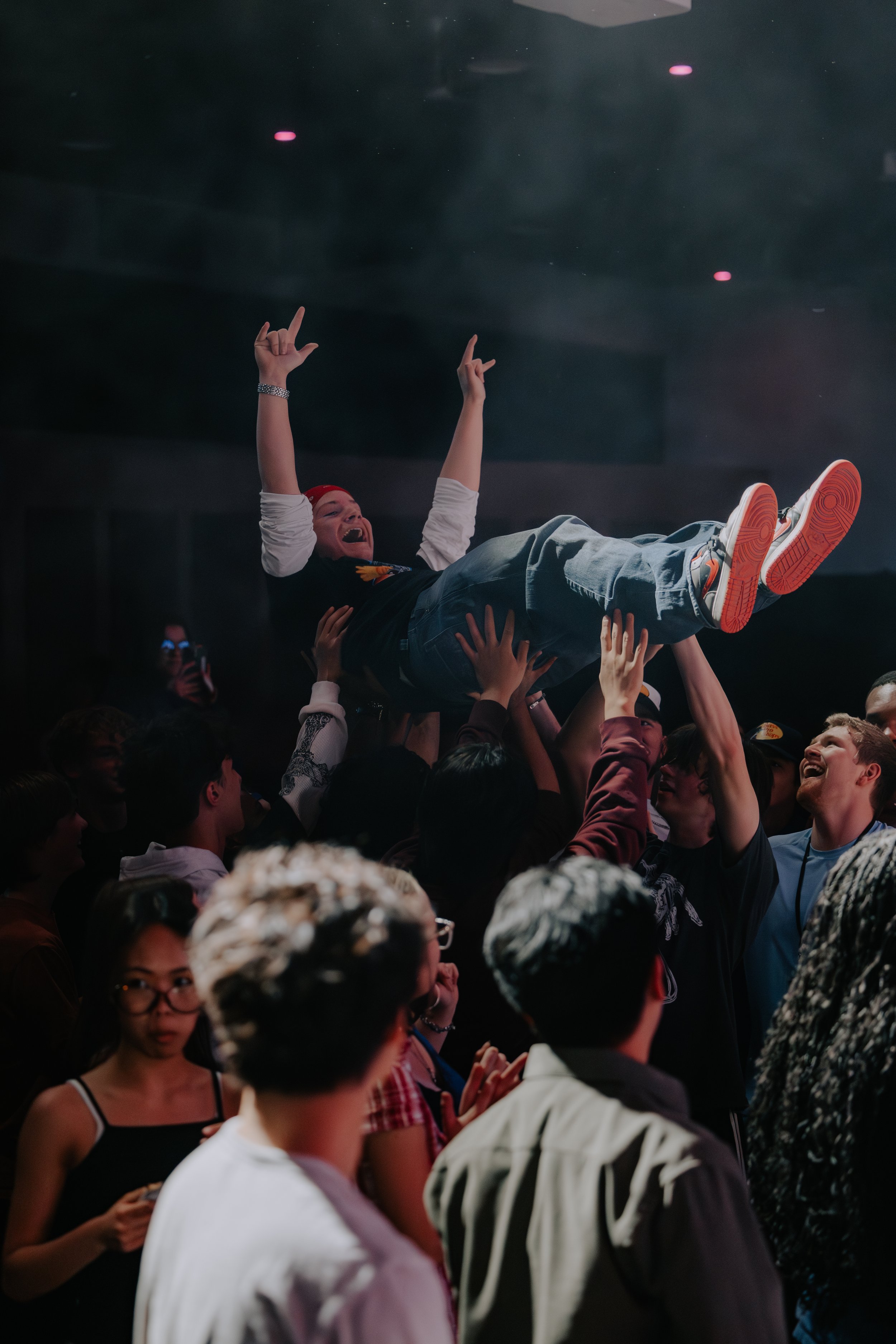 Person being lifted in the air by a crowd at a youth event, smiling and gesturing with their hands, surrounded by a diverse group of people.