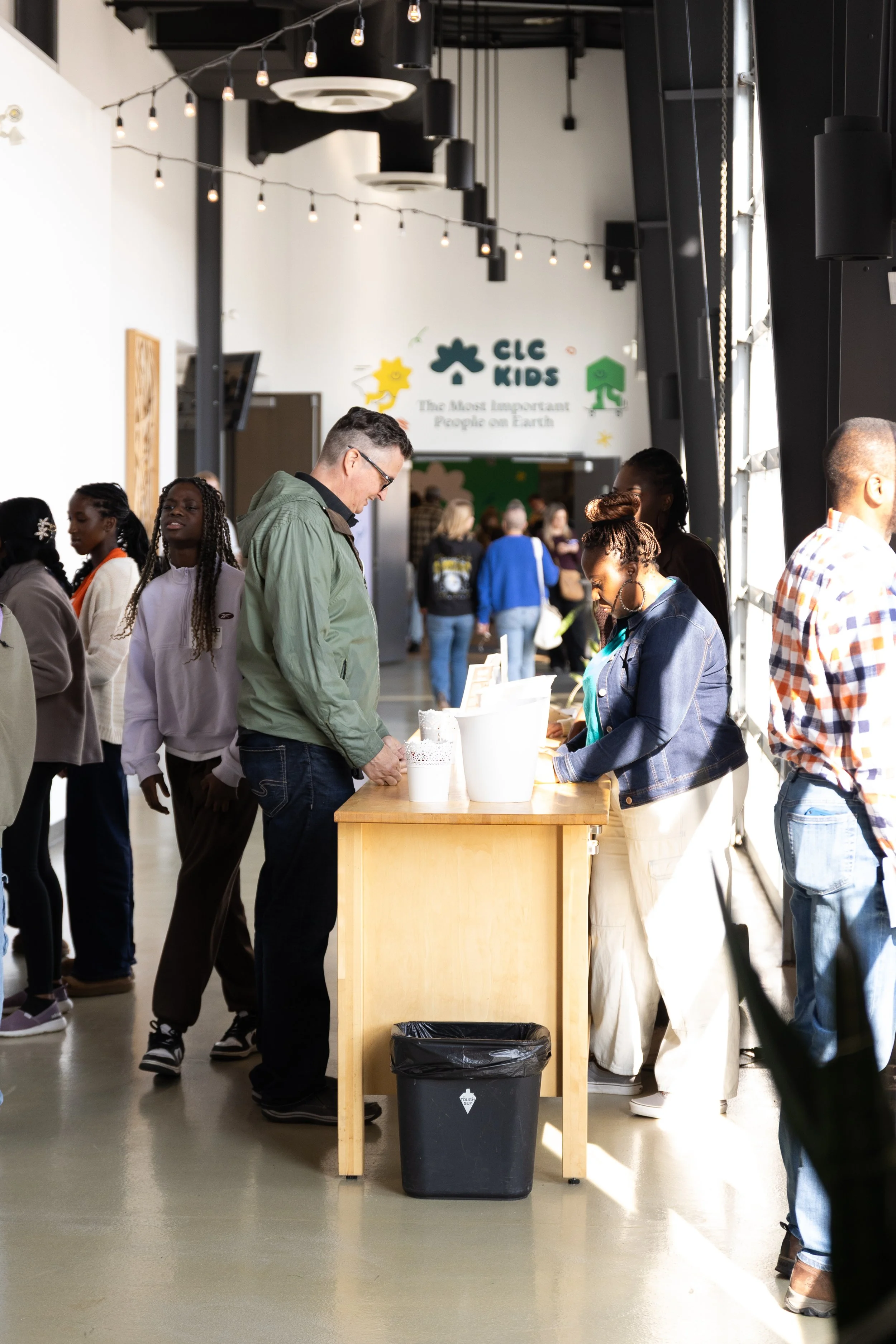 People standing in line at a registration or check-in desk inside a modern building with natural light. There is a sign in the background that reads "CLC KIDS" with a slogan underneath, and a table with a small white container and a black trash bin i