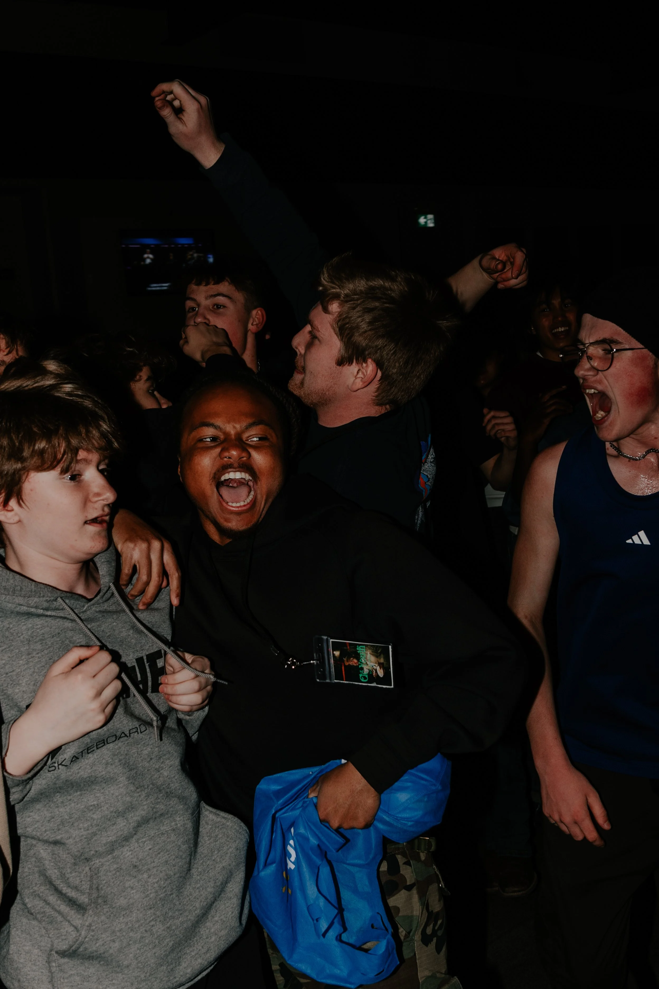 Group of people, mostly young men and women, celebrating with excitement and joy in a dark indoor setting. Some are smiling, shouting, and raising their fists.