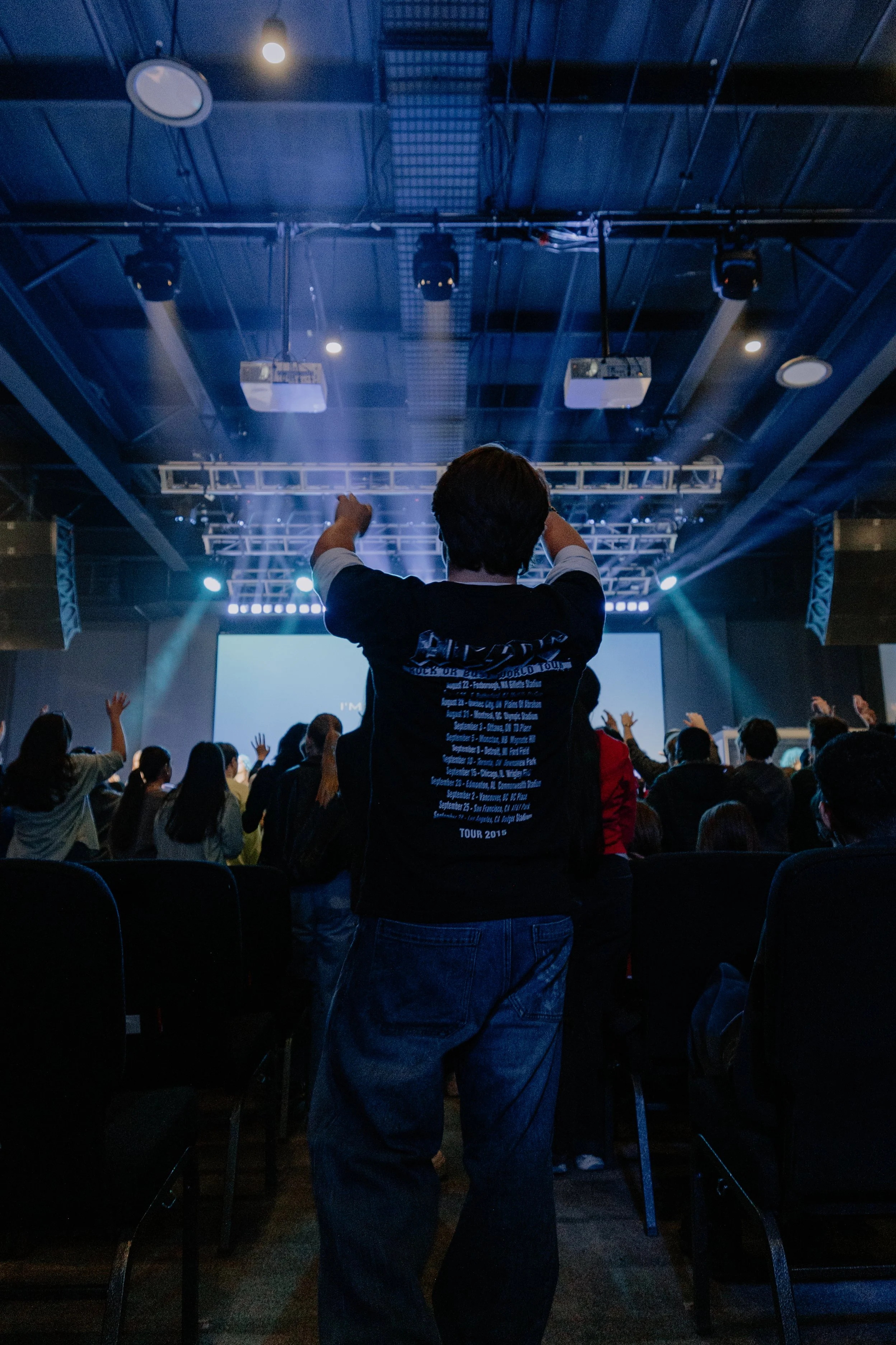 A person standing facing a stage with their hands raised, at a youthr event, in a large indoor venue with a crowd, stage lights, and a large screen displaying text in the background.