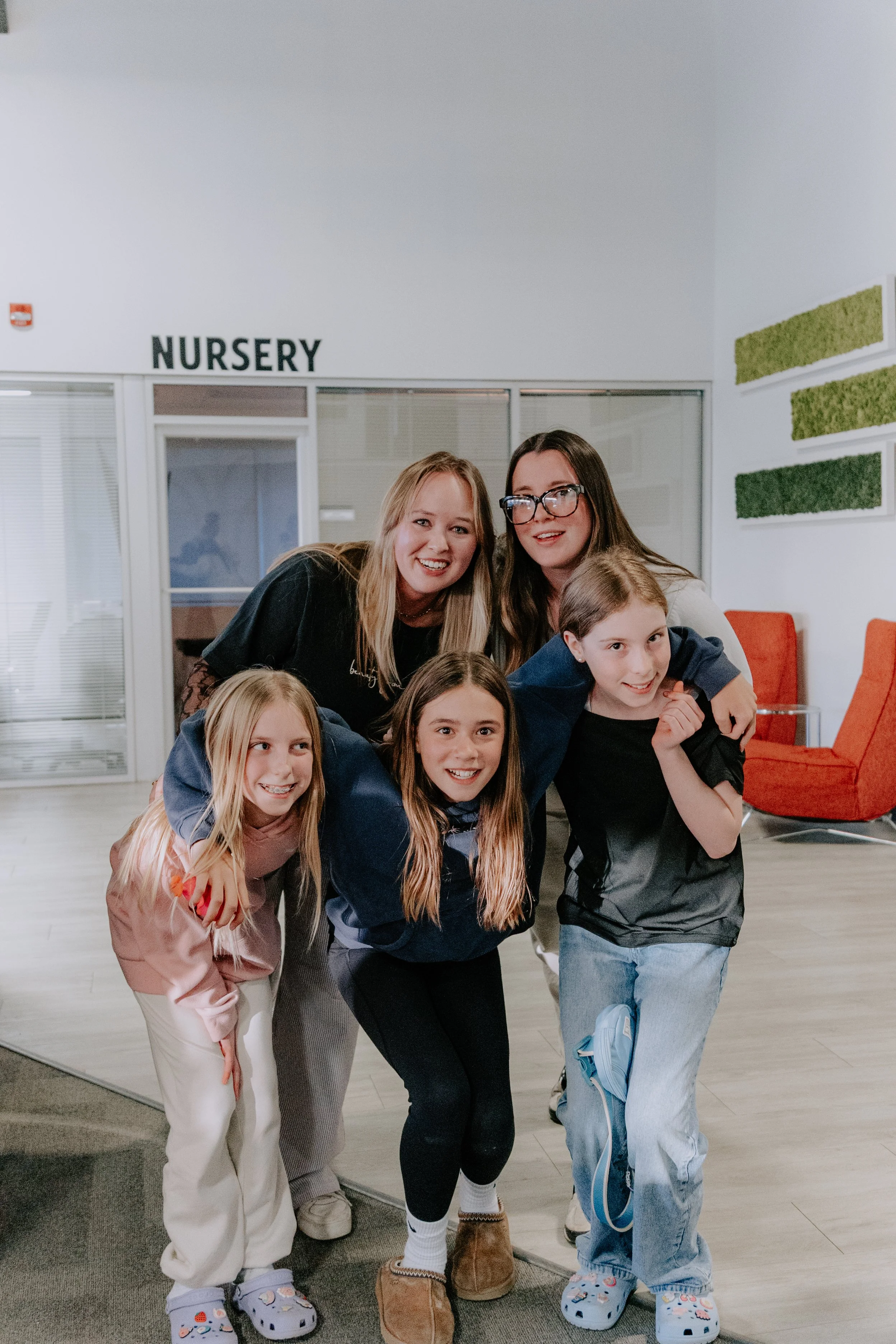 Group of children and two women smiling and posing indoors near a nursery sign, with some children wearing colorful shoes and casual clothing.