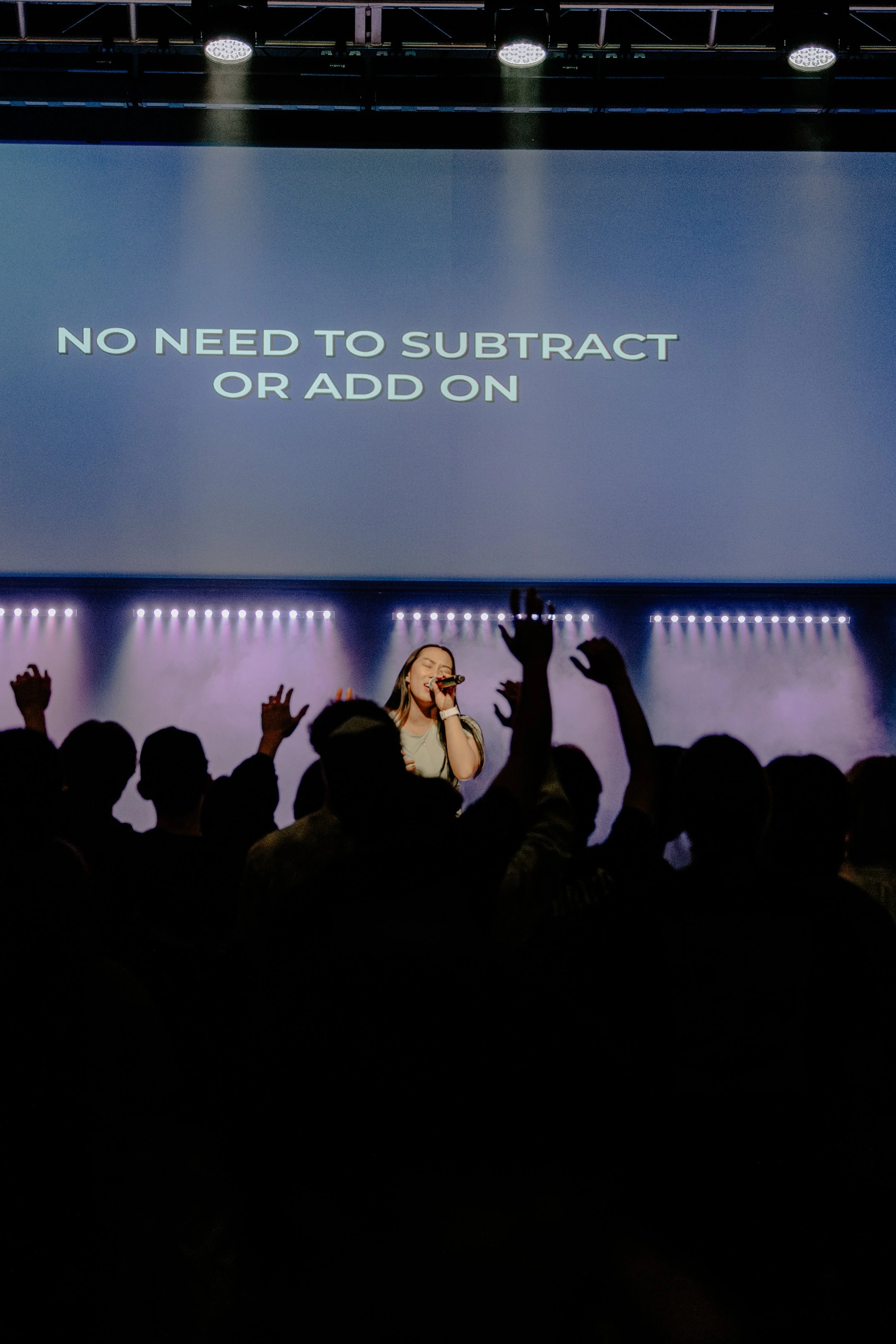 A concert scene with a singer on stage, holding a microphone, and an audience with raised hands in the foreground. A large screen behind displays the message, "NO NEED TO SUBTRACT OR ADD ON." The stage is illuminated with spotlights and colorful ligh