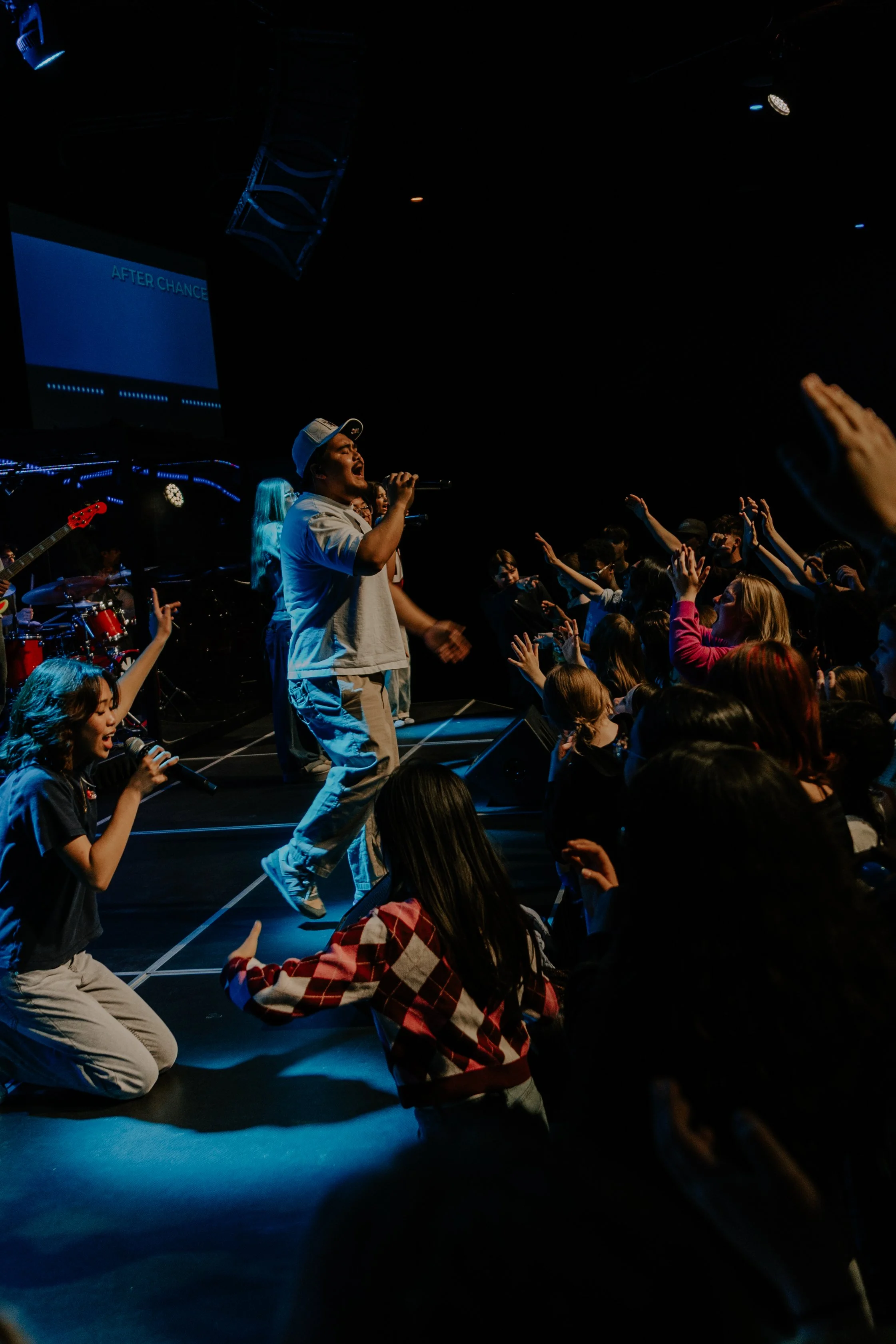 A man singing with a microphone on stage at a concert, surrounded by audience members reaching towards him with their hands in the air.