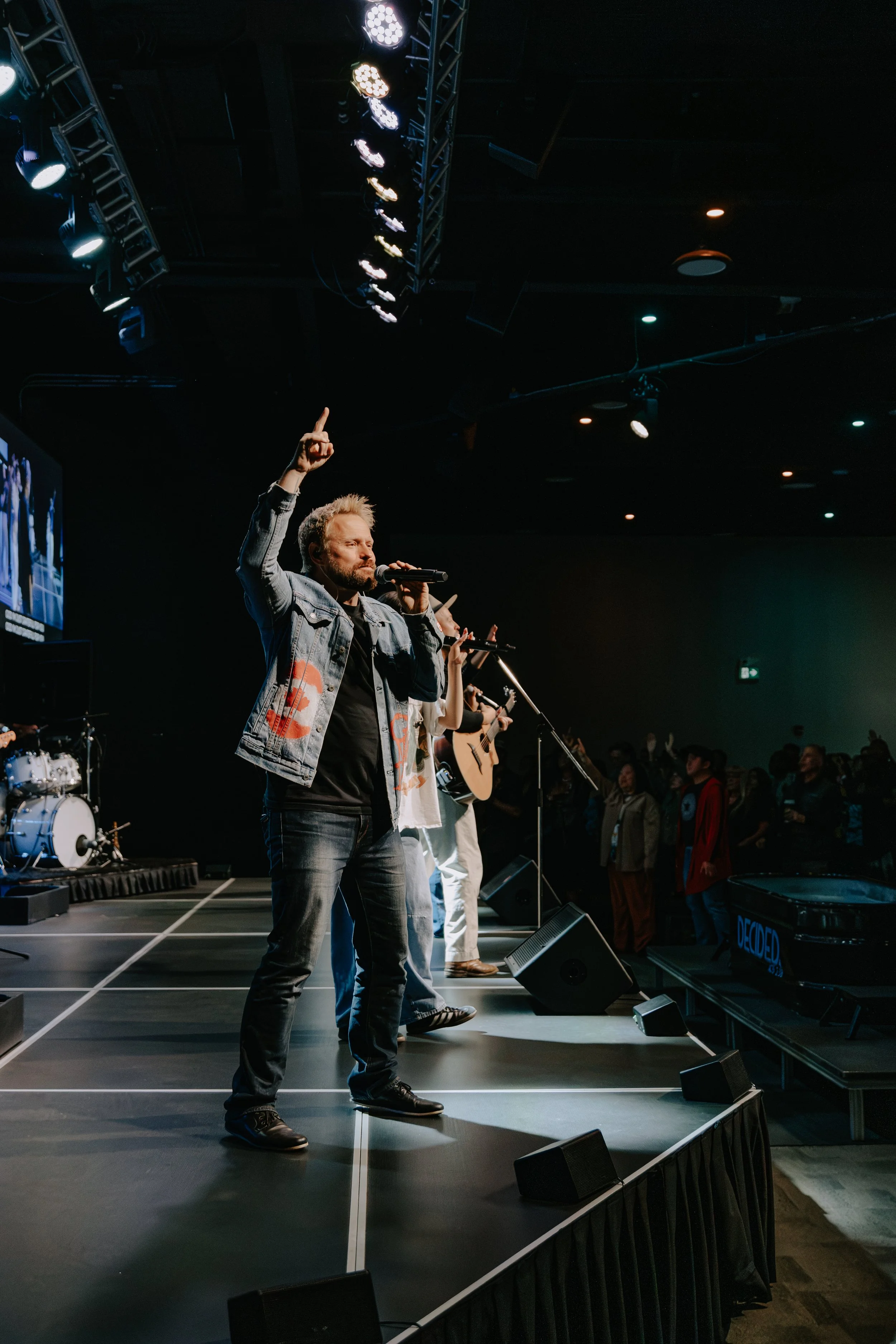 A man with a beard and light hair, wearing a denim jacket and black shoes, is standing on a stage, holding a microphone in his right hand and raising his left index finger. Behind him, there are several people with microphones, guitars, and drums, an