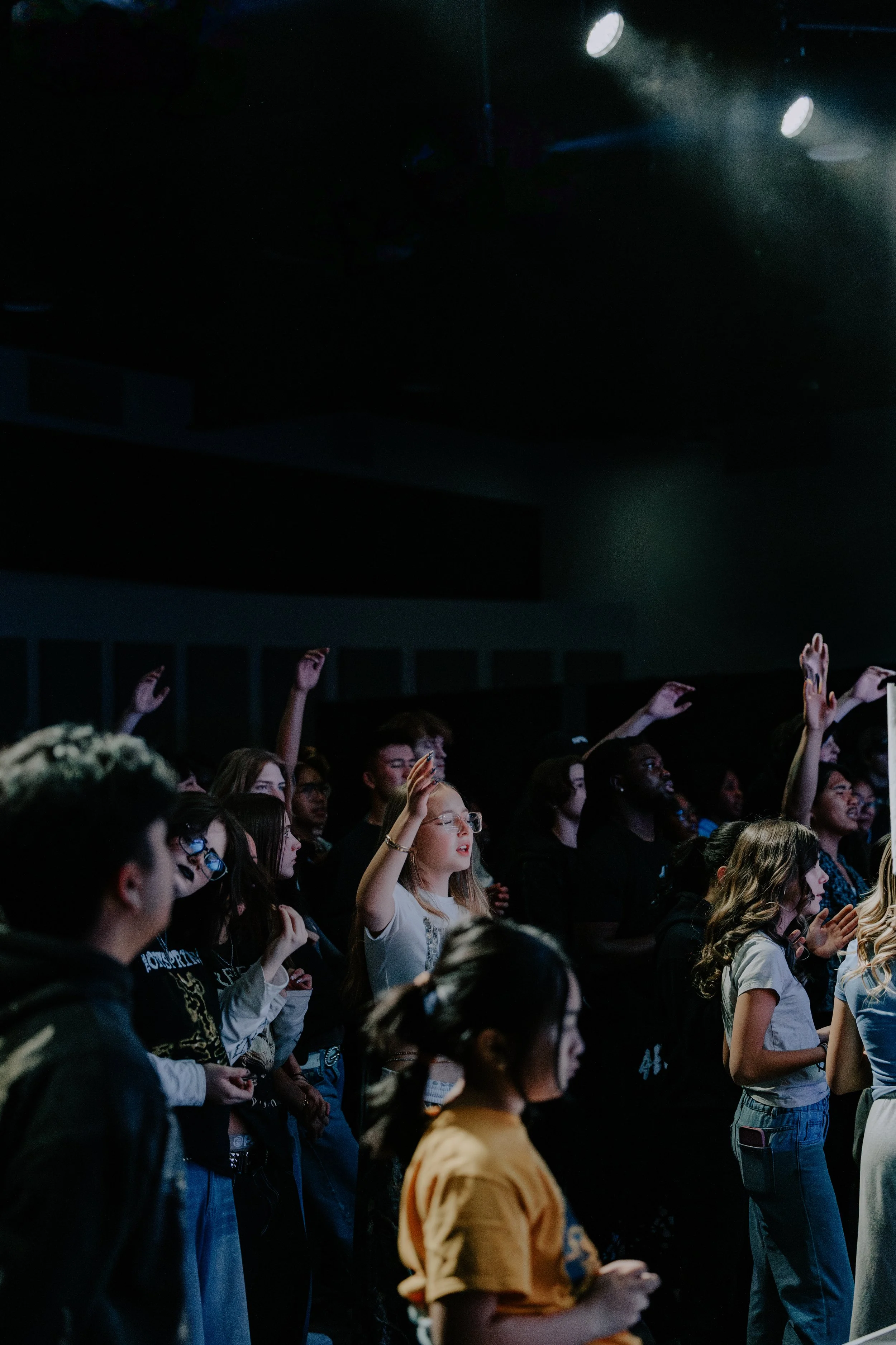 Audience members at a youth event raising their hands and paying attention to the stage with spotlights illuminating the ceiling.