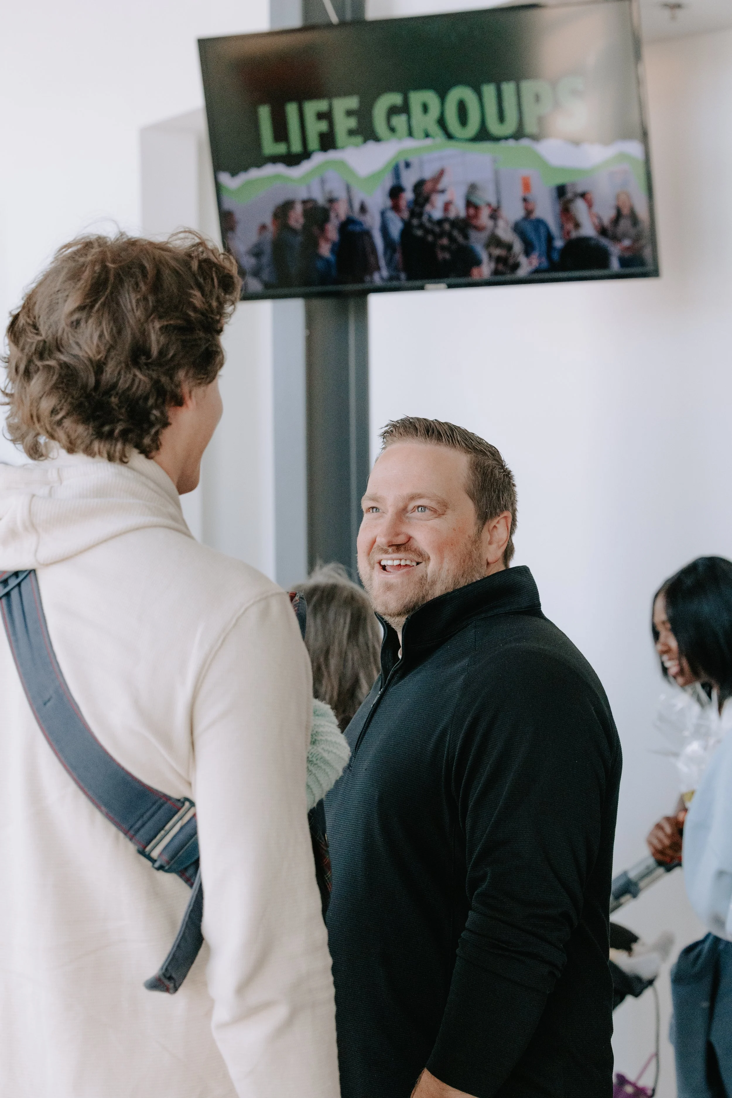 Two men are talking in a crowded room with a large screen on the wall displaying 'LIFE GROUP' and a group photo. One man has curly hair and a light-colored hoodie, the other has short hair and is wearing a black jacket, smiling and facing each other.