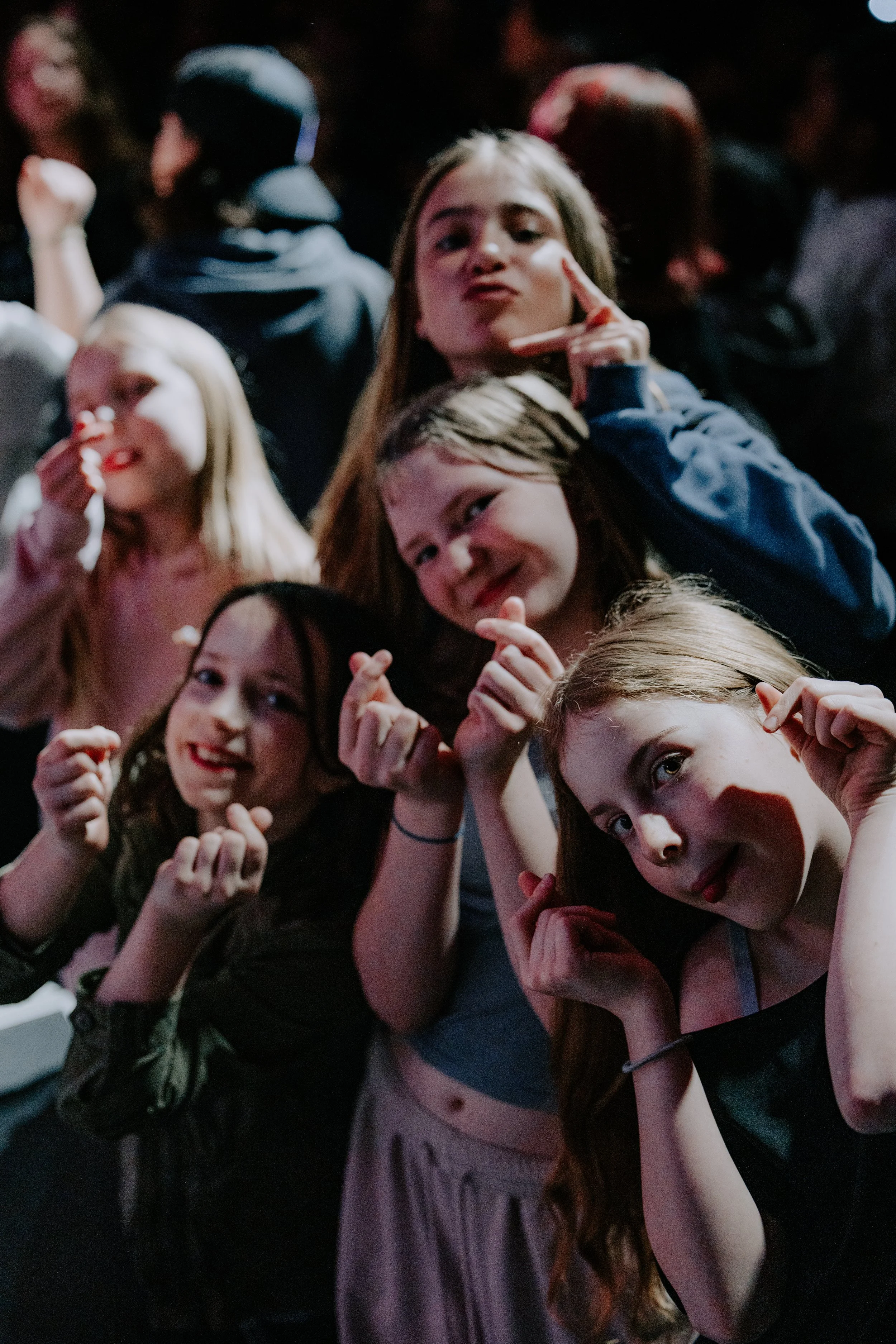 Group of young women posing together at a crowded event, making finger heart gestures and smiling at the camera.