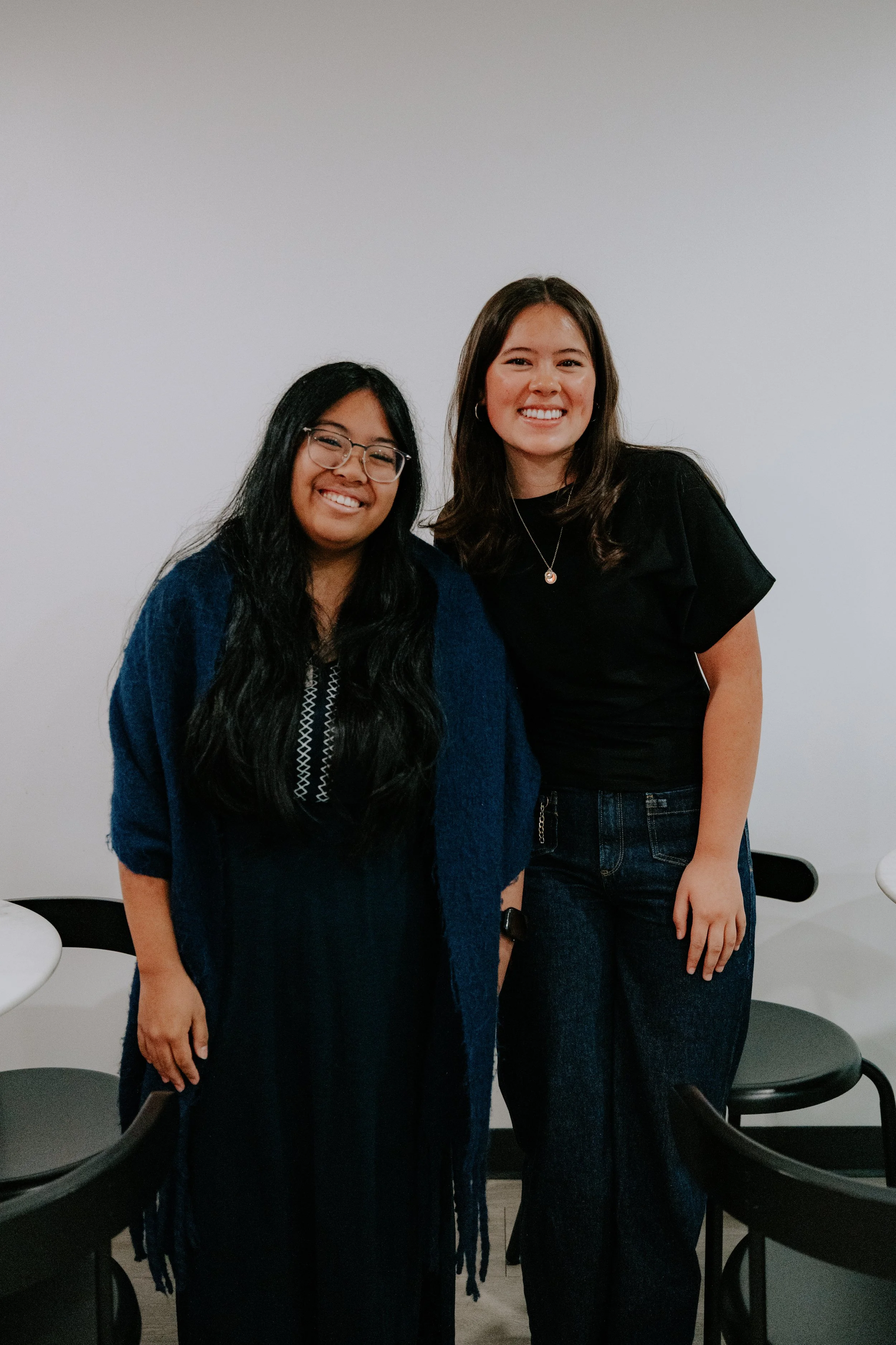 Two women standing together indoors, smiling, with chairs and a plain white wall in the background.