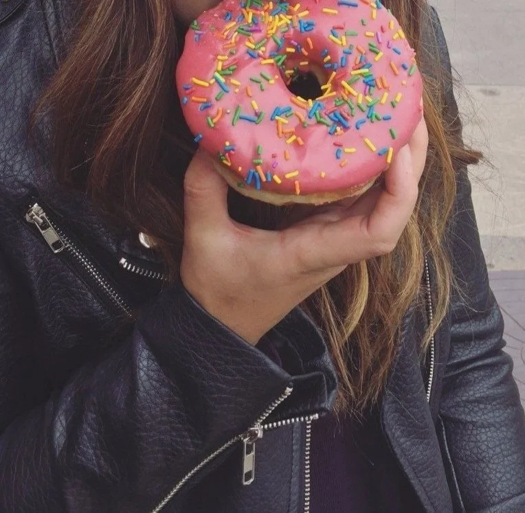 Person holding a donut with pink icing and colorful sprinkles, wearing a black leather jacket, with long brown hair.