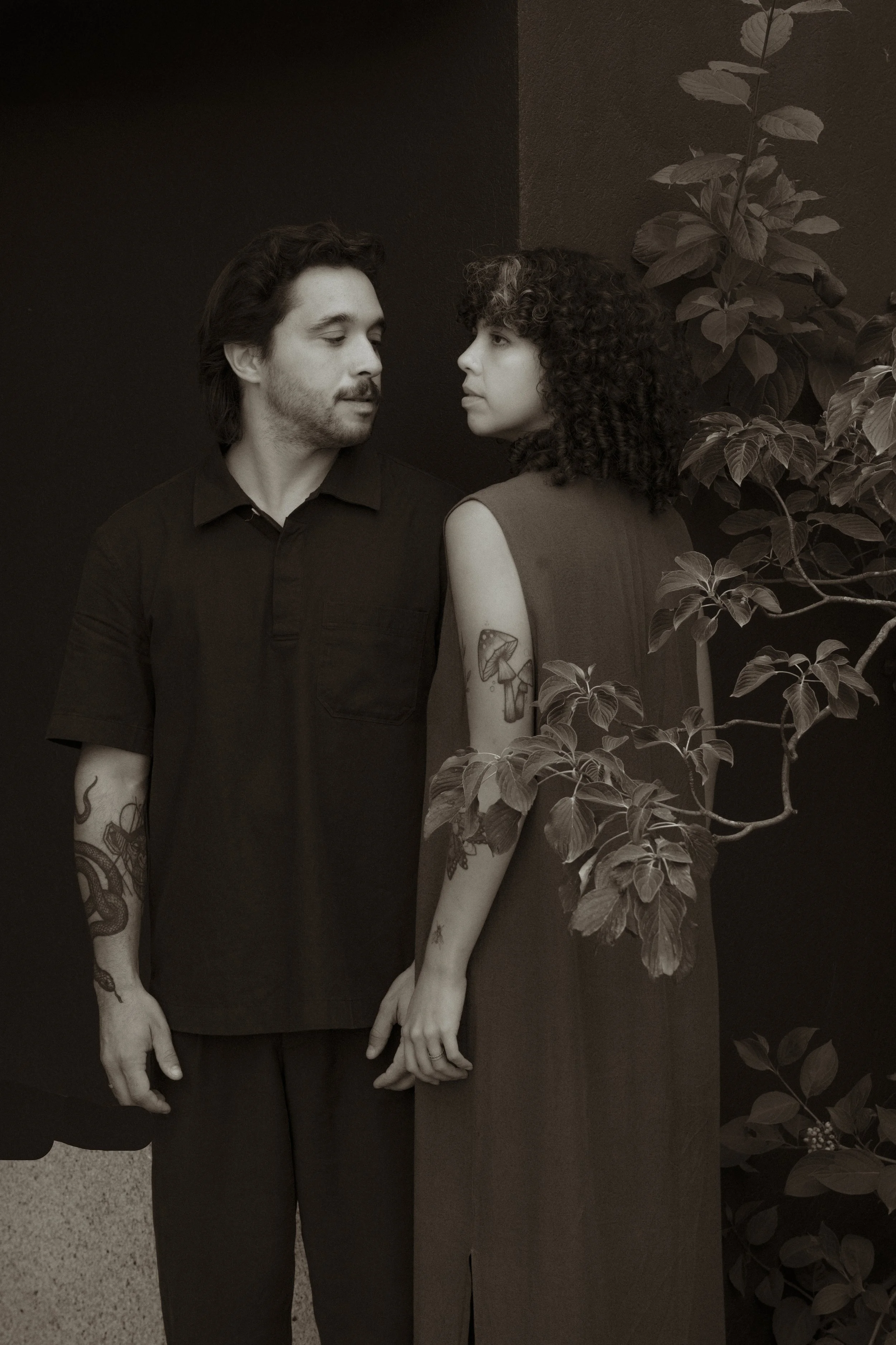 A man and woman holding hands, standing close together against a dark background with some leafy plants, looking at each other.