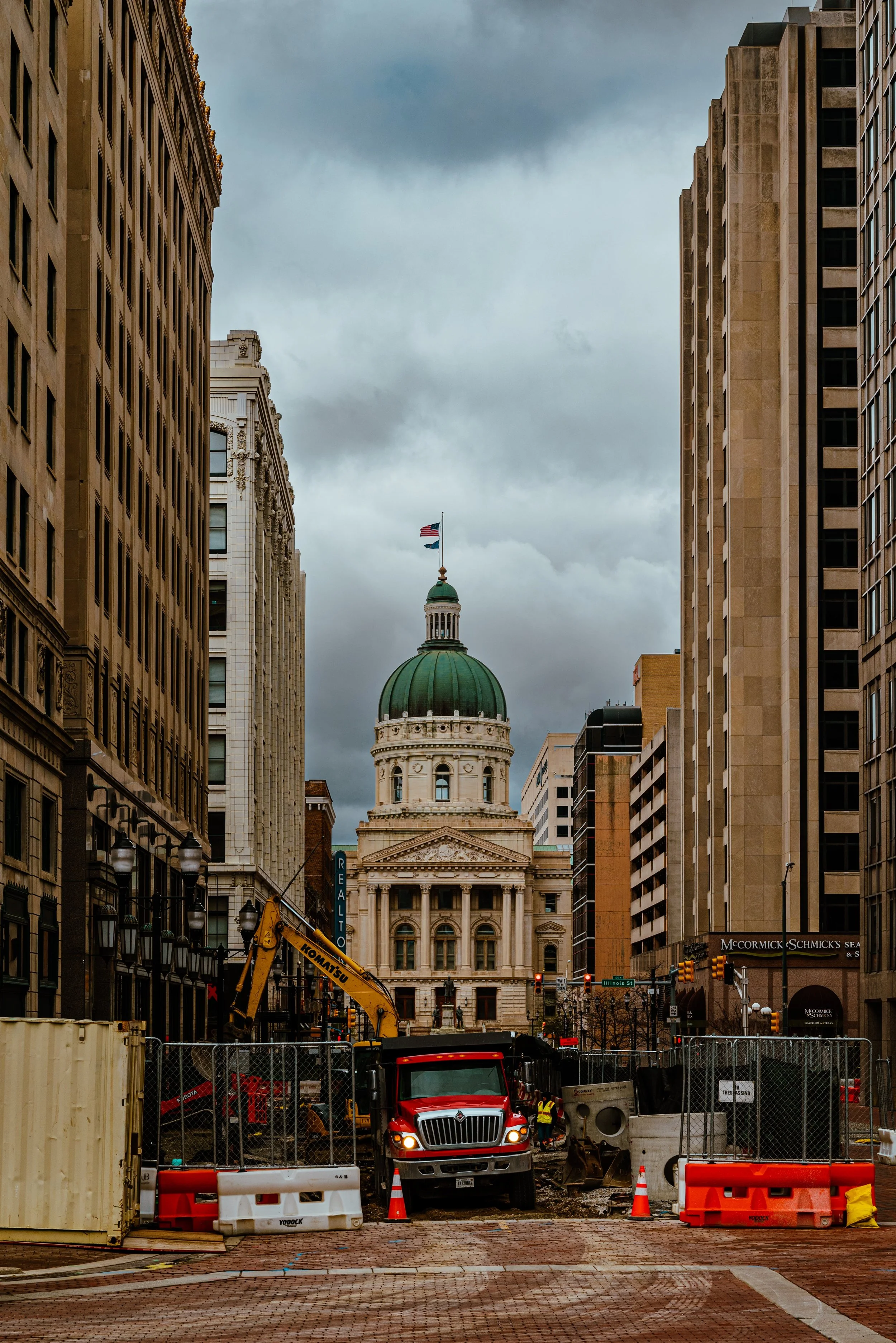 Downtown city street with historical government building, construction equipment, and barriers under cloudy sky.