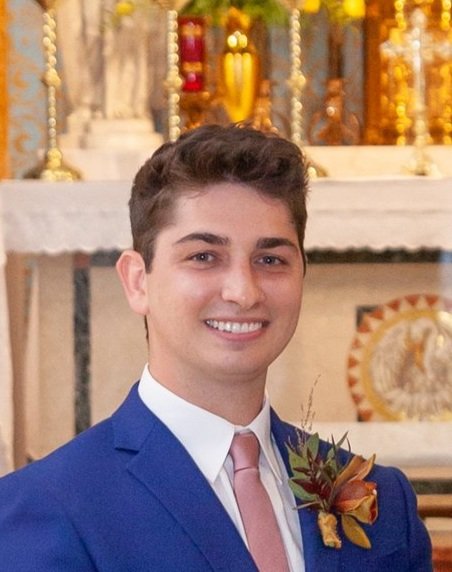Young man in a blue suit with a pink tie and a floral boutonniere, smiling at a celebration or ceremony with a decorated background.