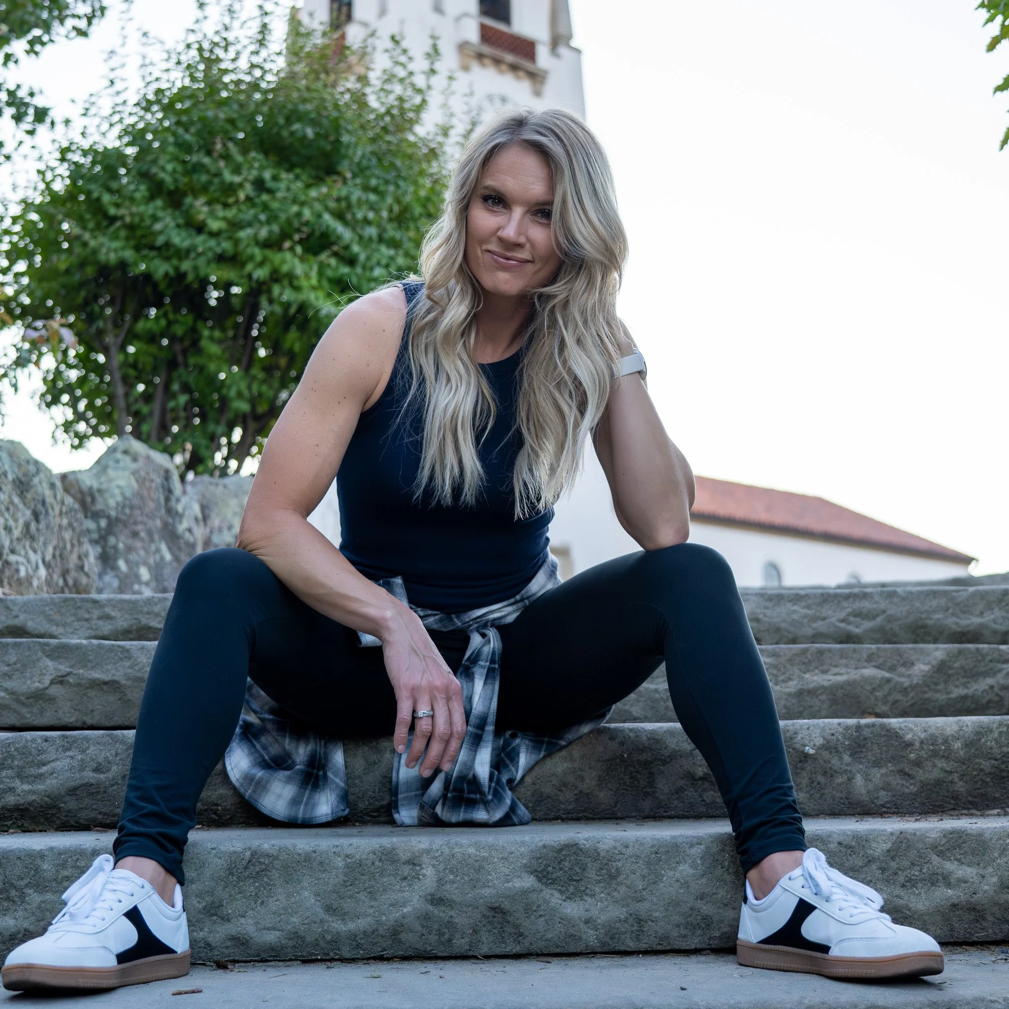 A woman with long blonde hair, wearing a navy tank top, black leggings, white sneakers, and a plaid shirt tied around her waist, sitting on outdoor steps with greenery and a building in the background.