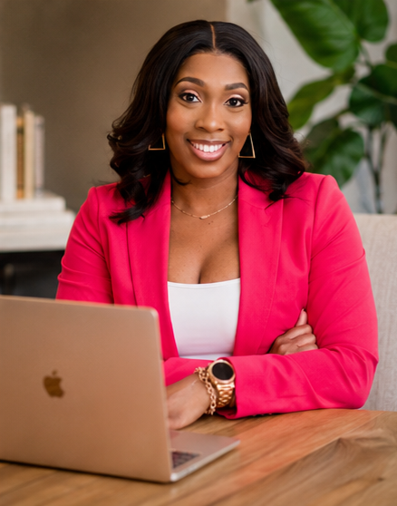 A woman sitting at a desk with a laptop, smiling at the camera, in a professional setting with a large green plant in the background.