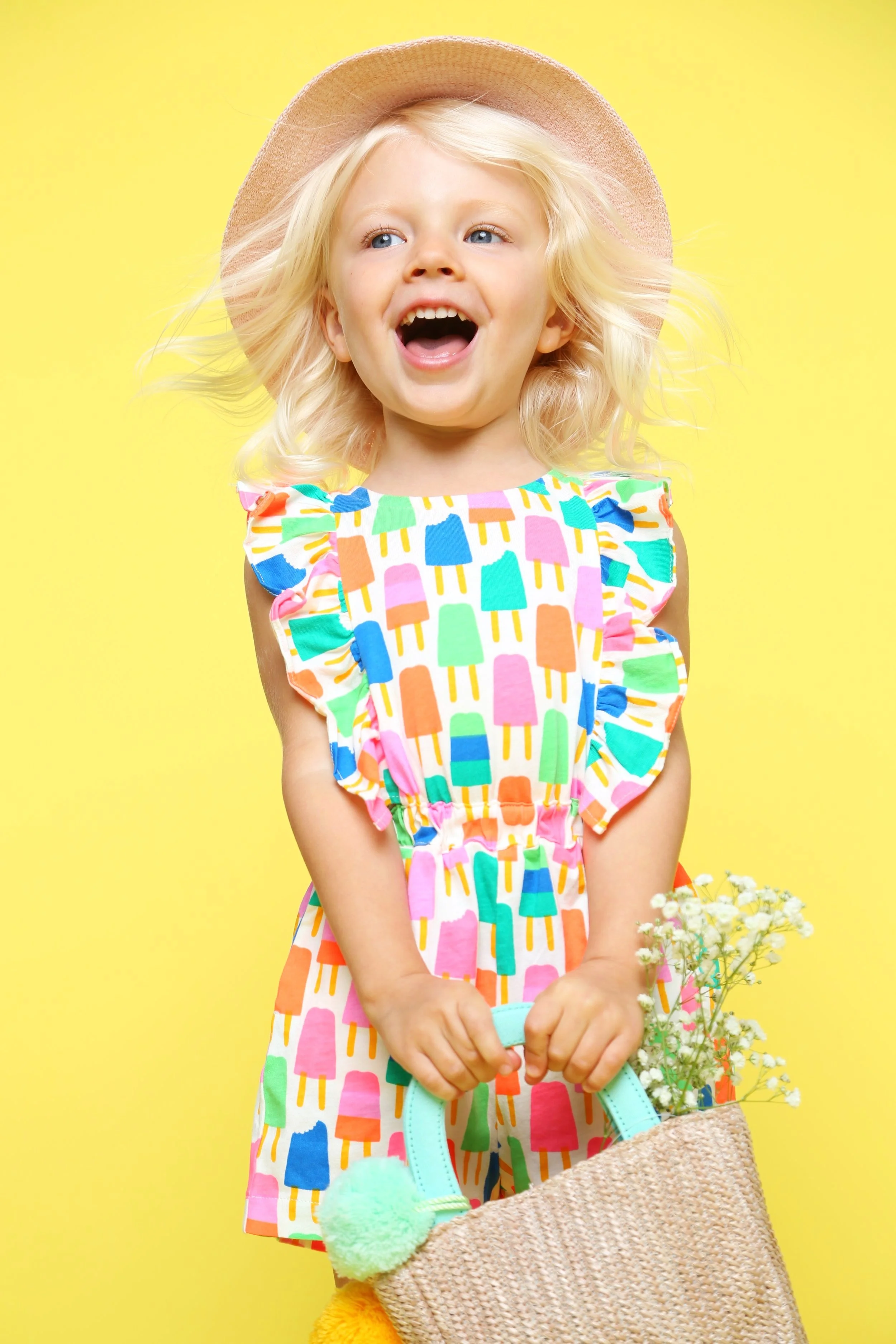 Child wearing a colorful popsicle dress and straw hat, holding a woven basket with flowers, on a yellow background.