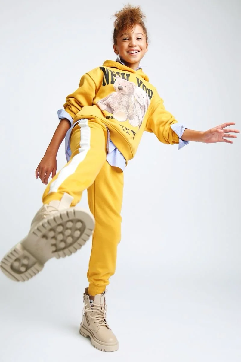 Child wearing yellow tracksuit with teddy bear graphic, beige boots, and blue shirt underneath, smiling with one leg raised.